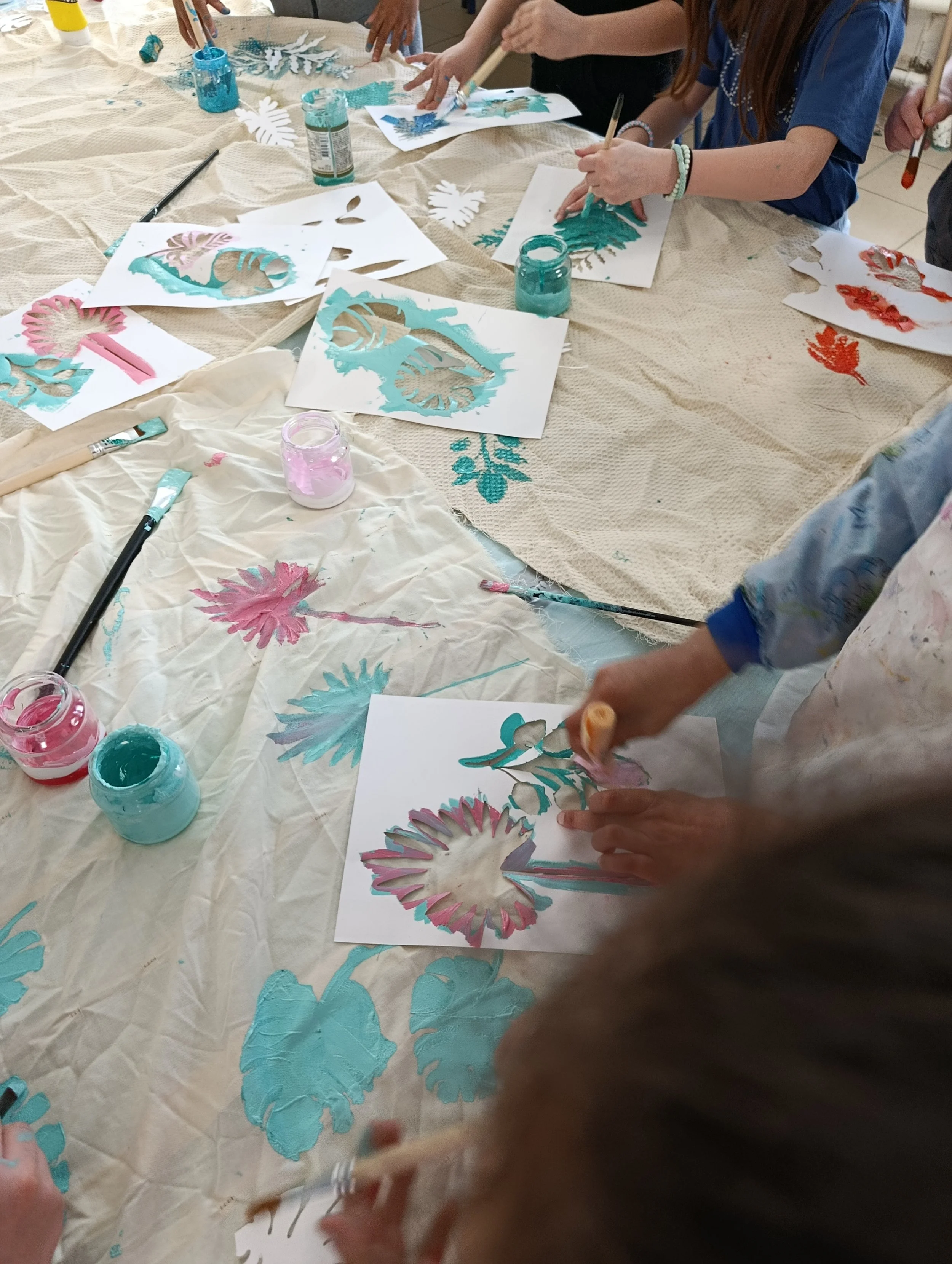 Groupe d'enfants et d'adultes réalisant des œuvres d'art avec des pinceaux et des peinture sur papier, entourés de matériaux de bricolage colorés.