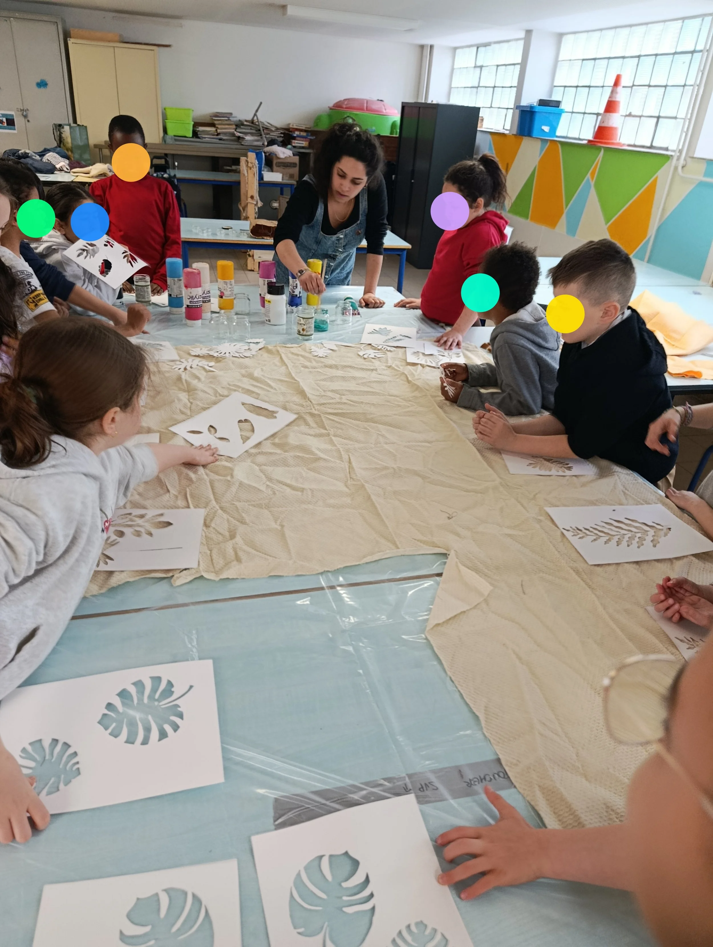 Une classe d'enfants en train de réaliser une activité artistique autour de découpes de feuilles tropicales en papier, sous la supervision d'une adulte.