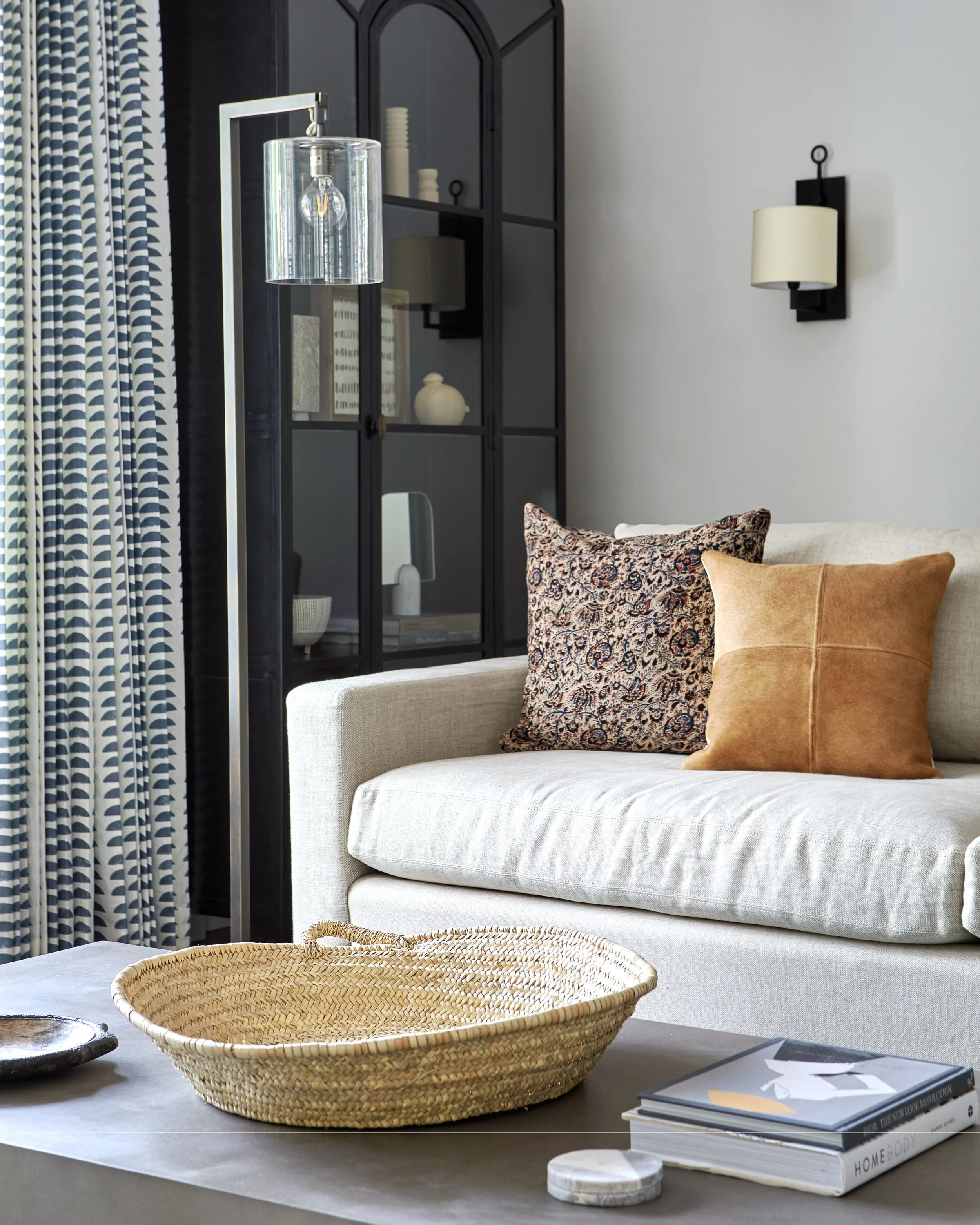 Living room with a beige sofa, two decorative pillows, a black cabinet, a floor lamp, a woven basket on the coffee table, and a stack of magazines.