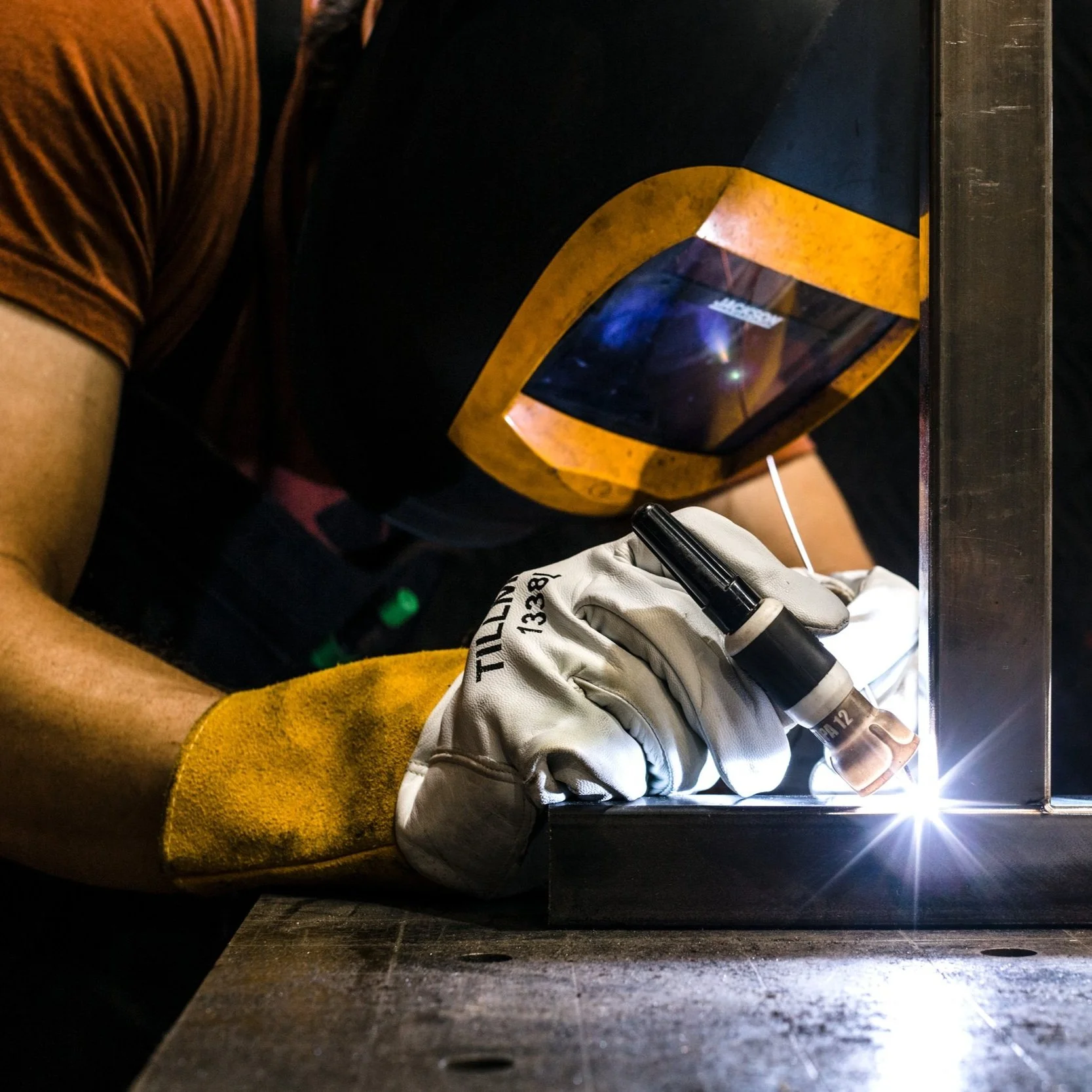 A welder wearing a protective helmet and gloves welding metal outdoor at night, sparks and bright light from the welding process.