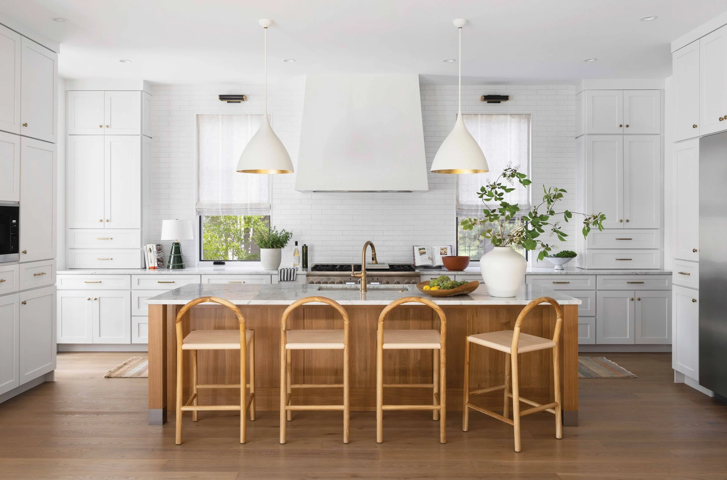 A modern white kitchen with a central island, four wooden chairs, pendant lights, and a white range hood, with windows letting in natural light and green plants.