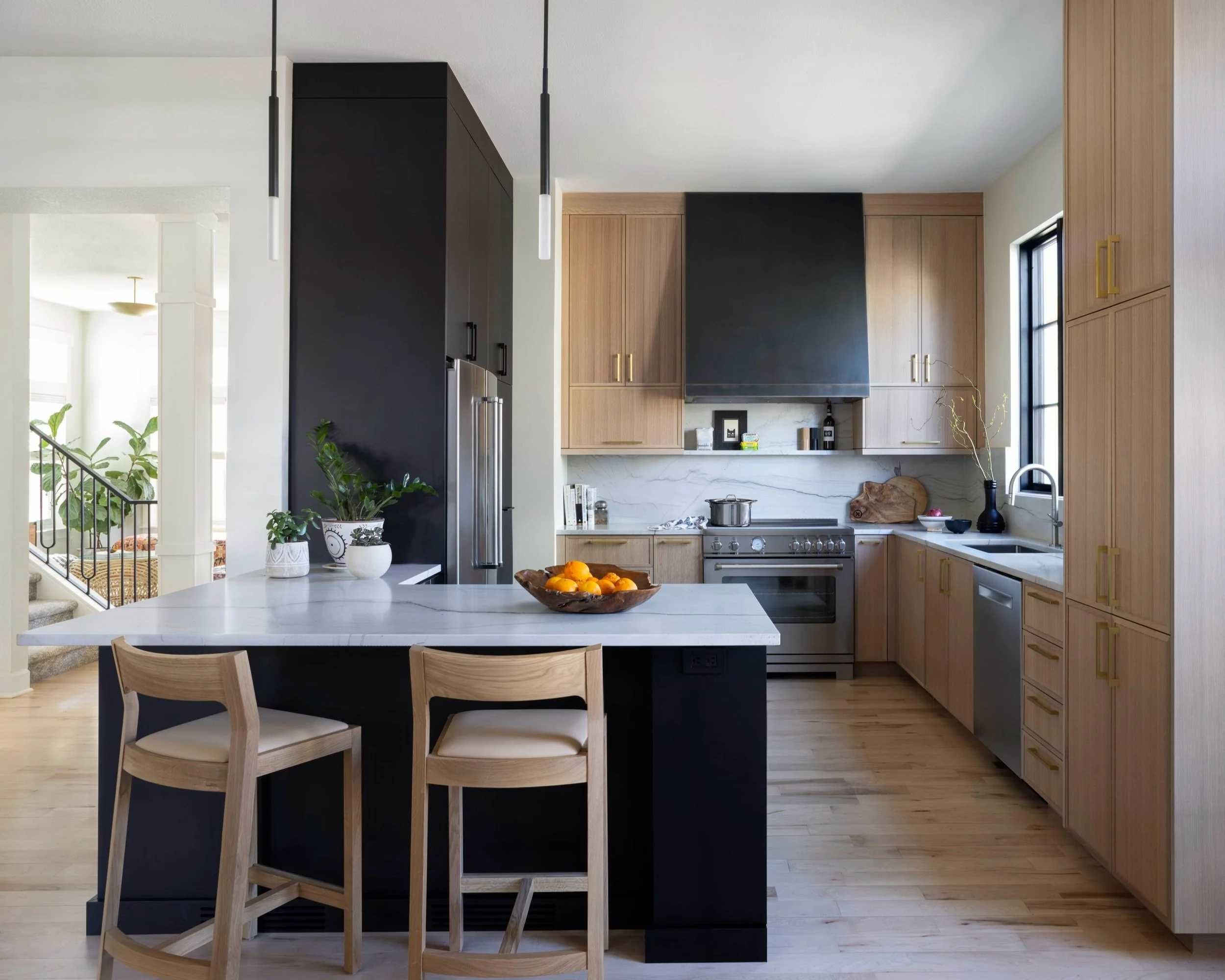 Modern kitchen with light wood cabinets, stainless steel appliances, black and white marble backsplash, central island with white countertop, two wooden stools, black and white pots with green plants, bowl of oranges, window with black frame, and black range hood.
