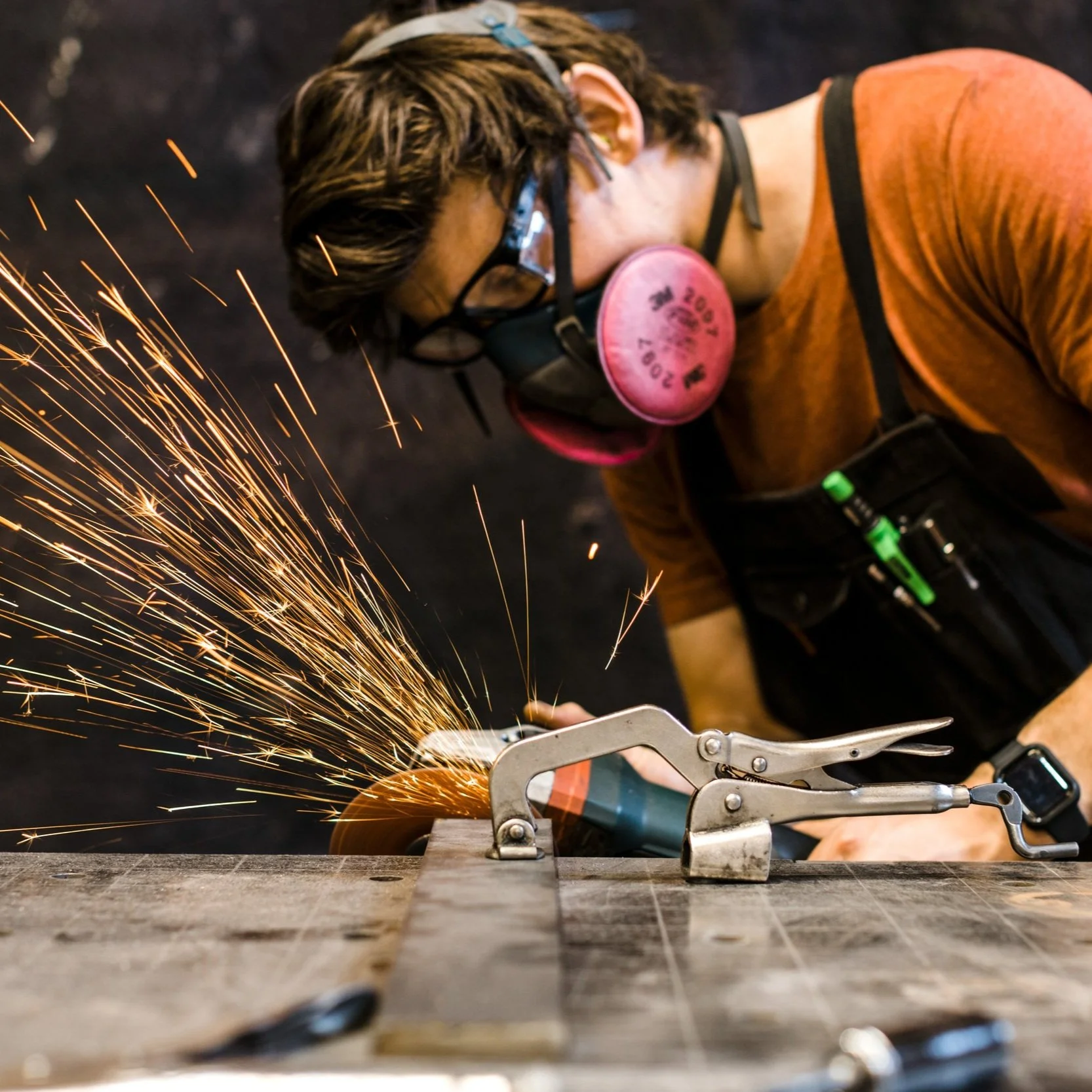 A person wearing safety glasses, a pink respirator, and a tool belt is grinding metal on a workbench, with sparks flying.
