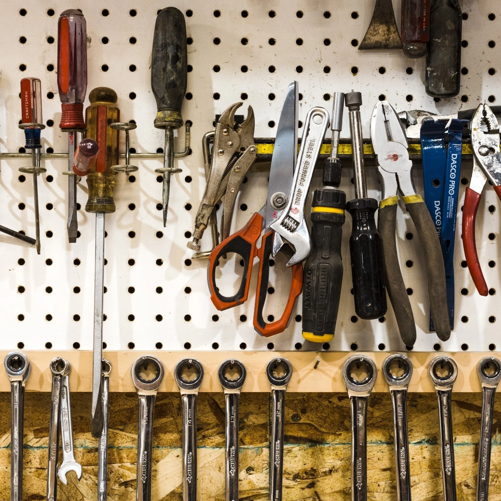 Tools hanging on a white pegboard, including screwdrivers, pliers, scissors, and a wrench, with sockets arranged below on a wooden strip.