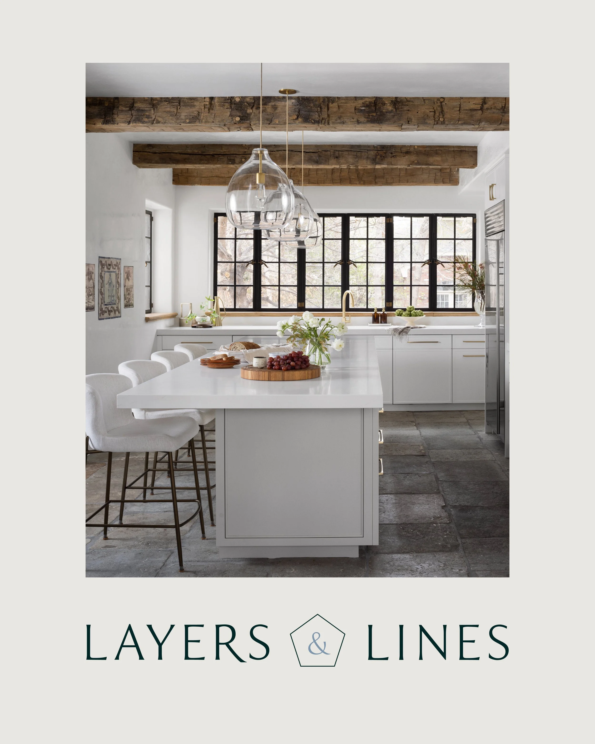 Modern white kitchen with wooden beams, large window, and a white island with bar stools, decorated with flowers and grapes.