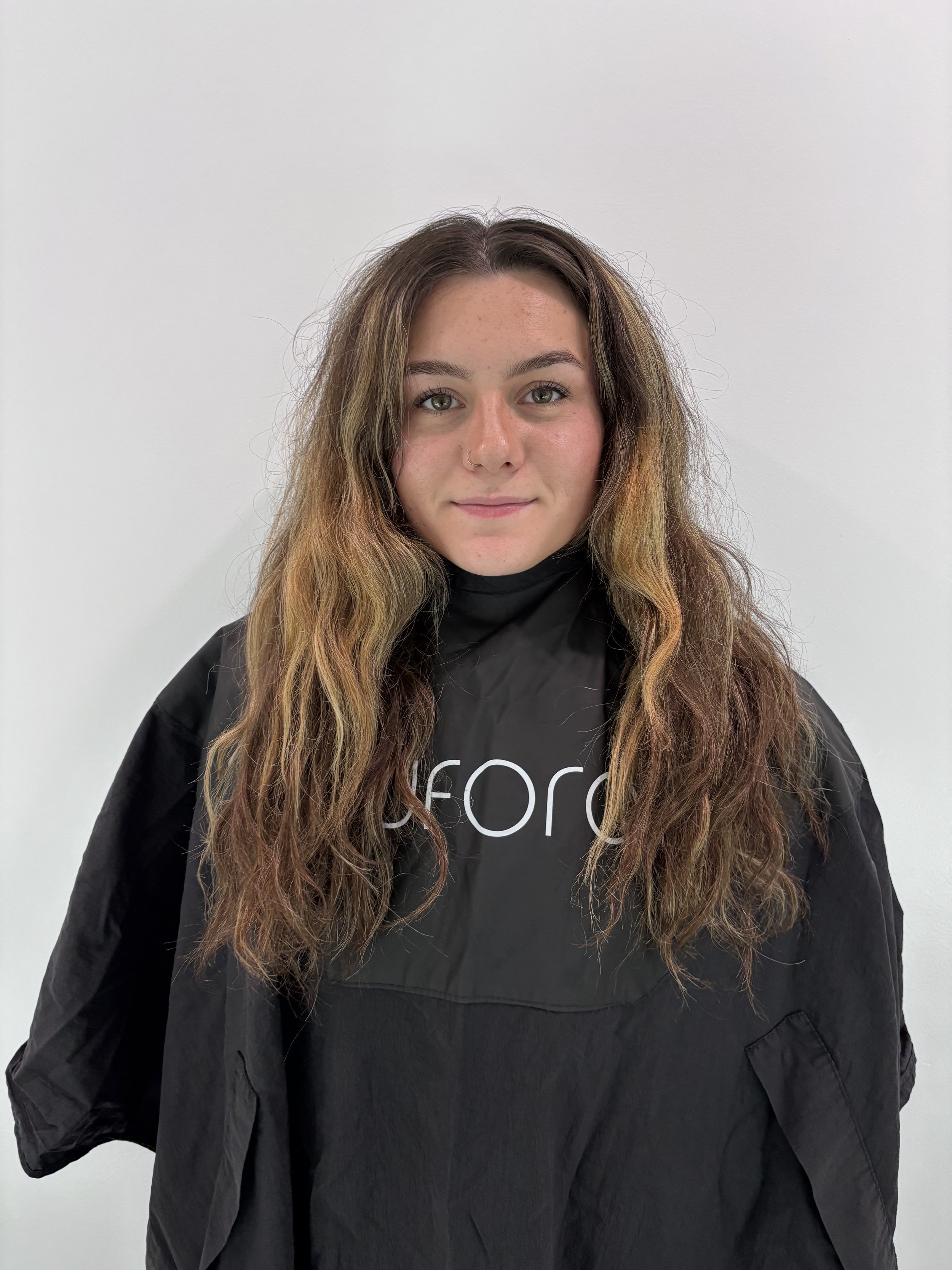 Young woman with long wavy hair, light complexion, and a nose piercing, wearing a black jacket and smiling softly in front of a plain white wall.