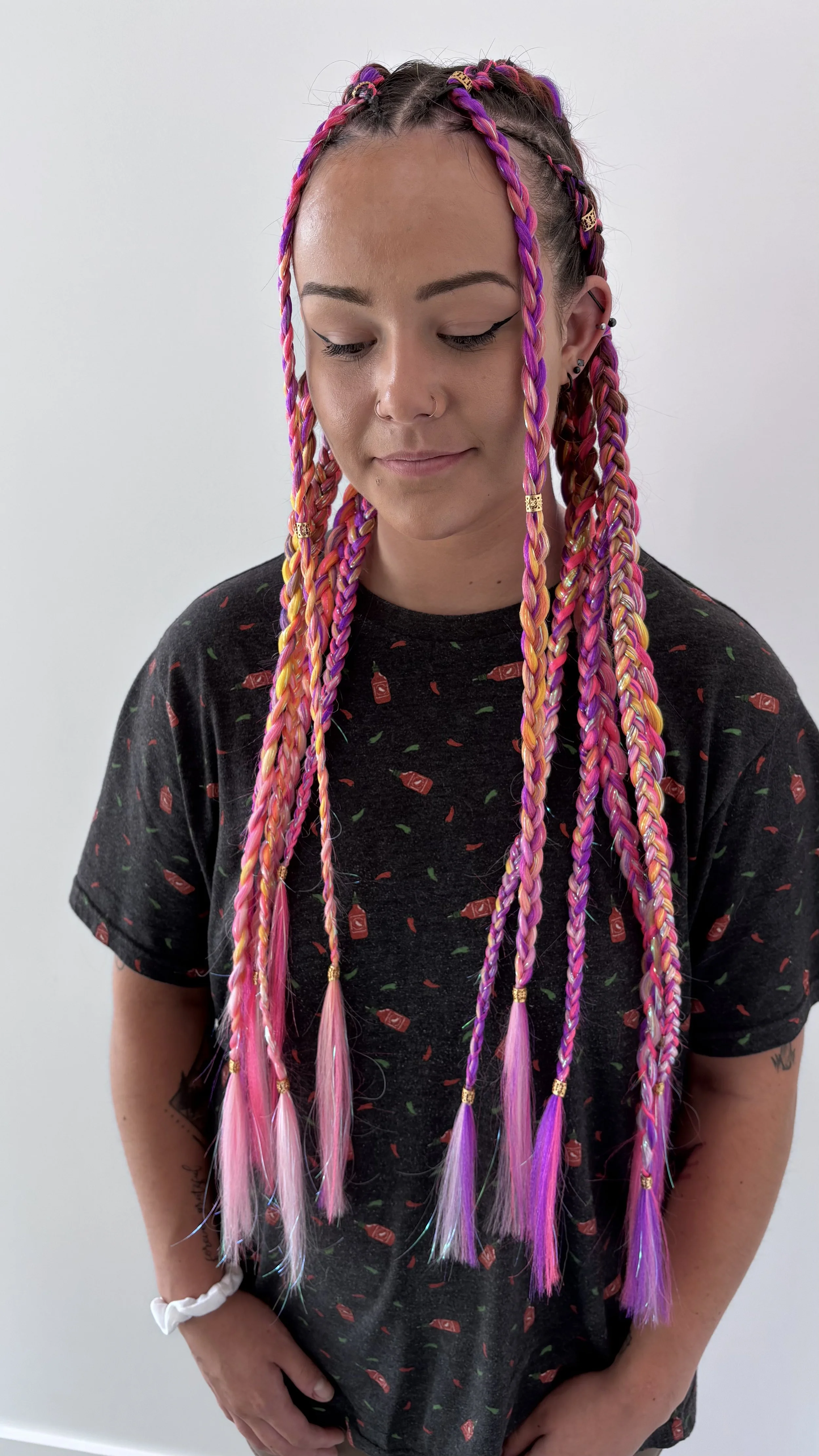 A young woman with colorful pink, purple, and blonde braided hair wearing a black t-shirt with hot sauce chili pepper prints, standing against a plain white background.