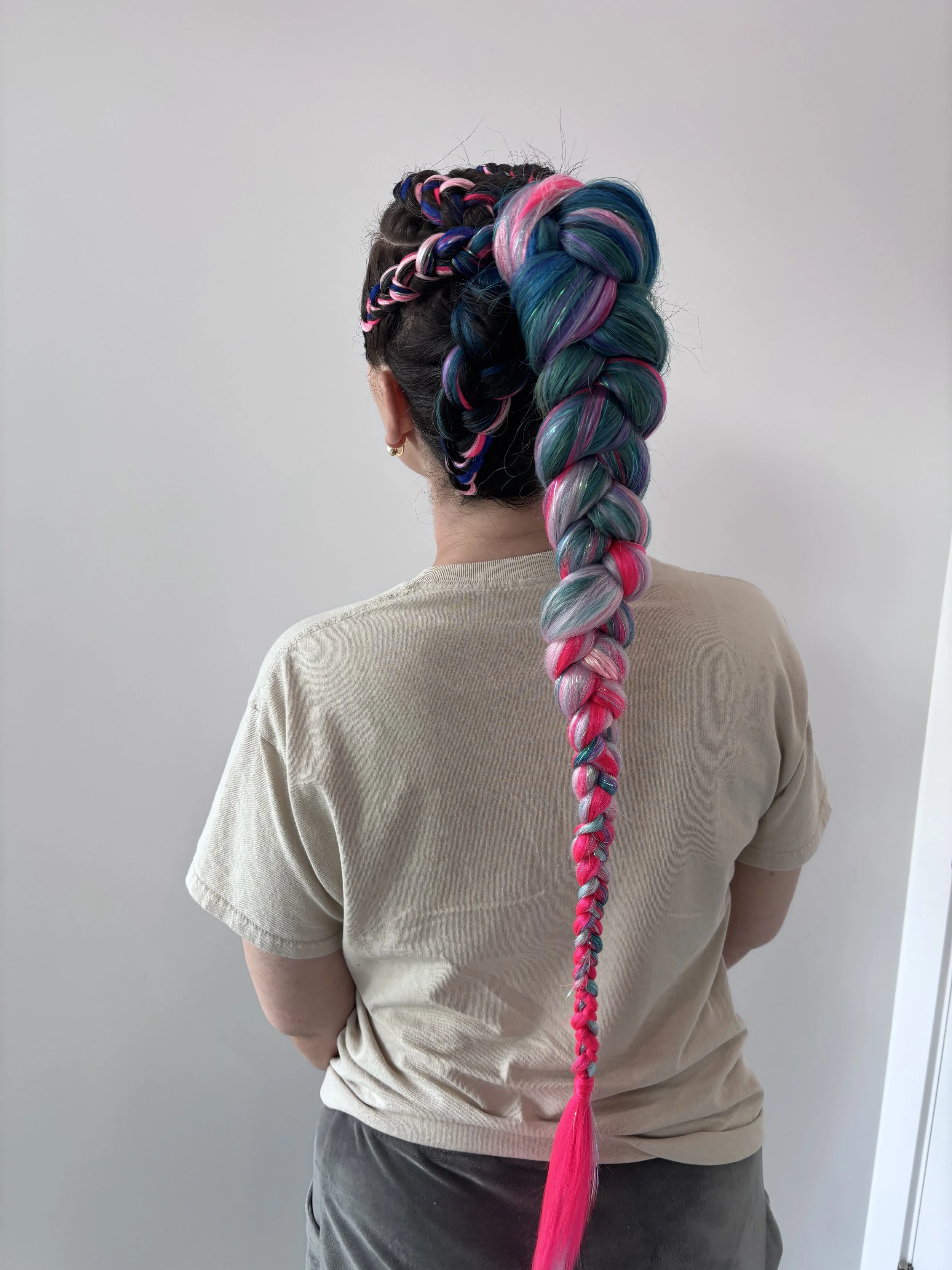 A person seen from the back with a colorful, multi-colored braided hairstyle featuring shades of pink, blue, purple, and green, standing against a plain, light-colored wall.