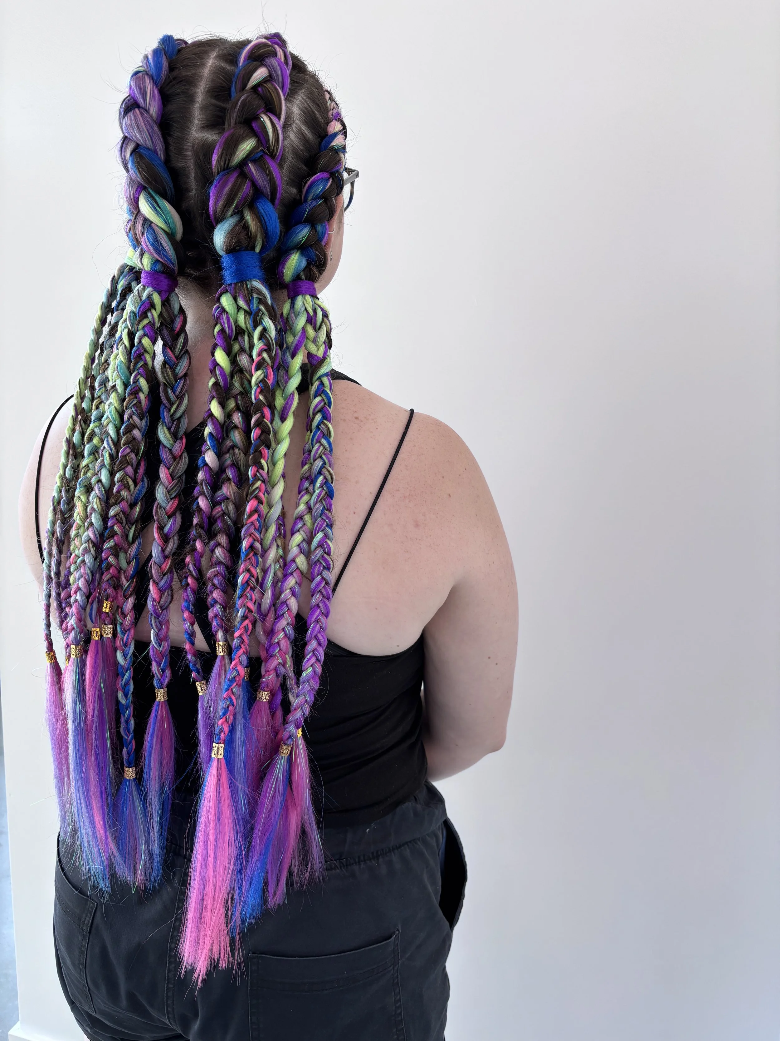 Back view of a woman with long multicolored braids, wearing glasses, black tank top, and black pants, against a plain white wall.