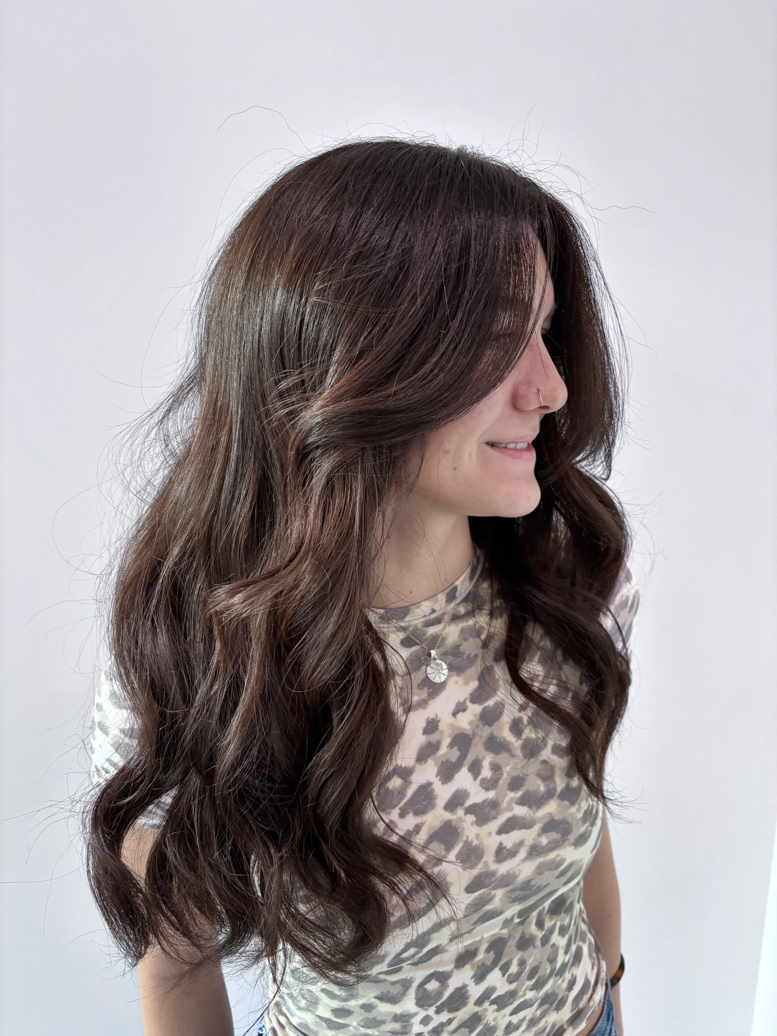 A woman with long, wavy brown hair with lighter highlights, wearing a leopard print top and a necklace, smiling and standing against a plain white background.