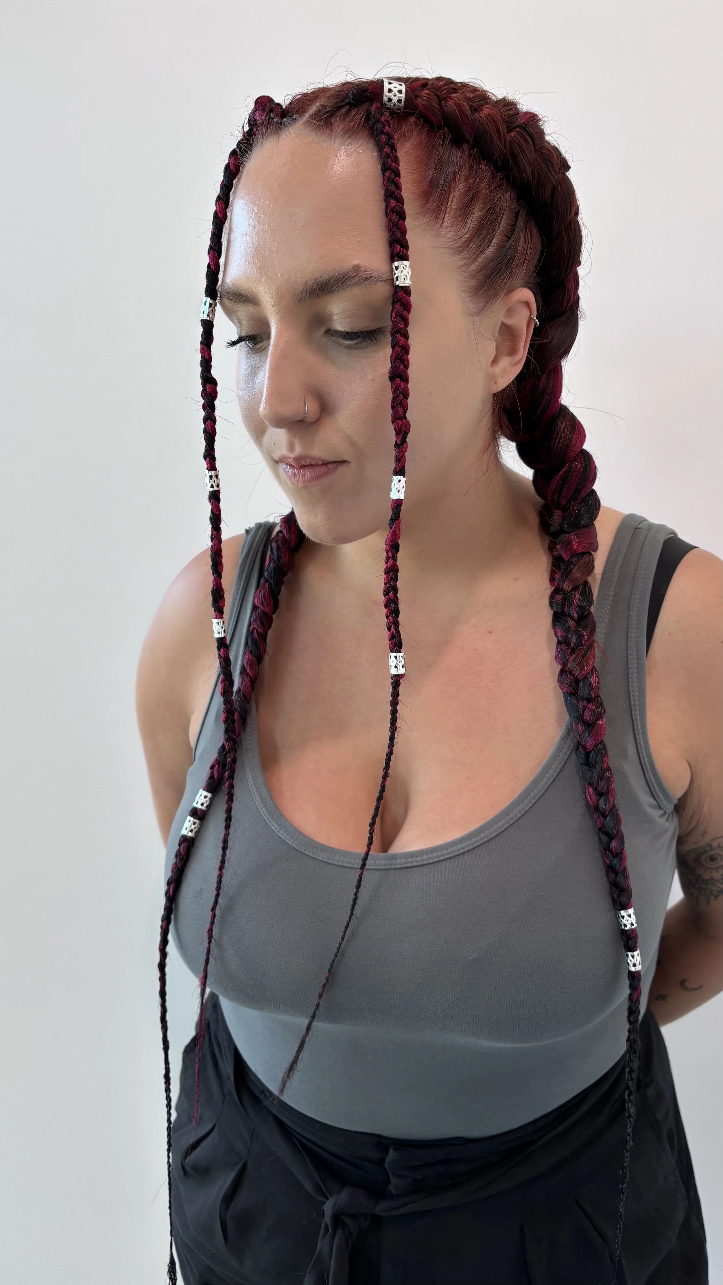 Woman with braided hair featuring black and red braids decorated with silver beads, wearing a gray tank top and black pants, standing against a plain white wall.