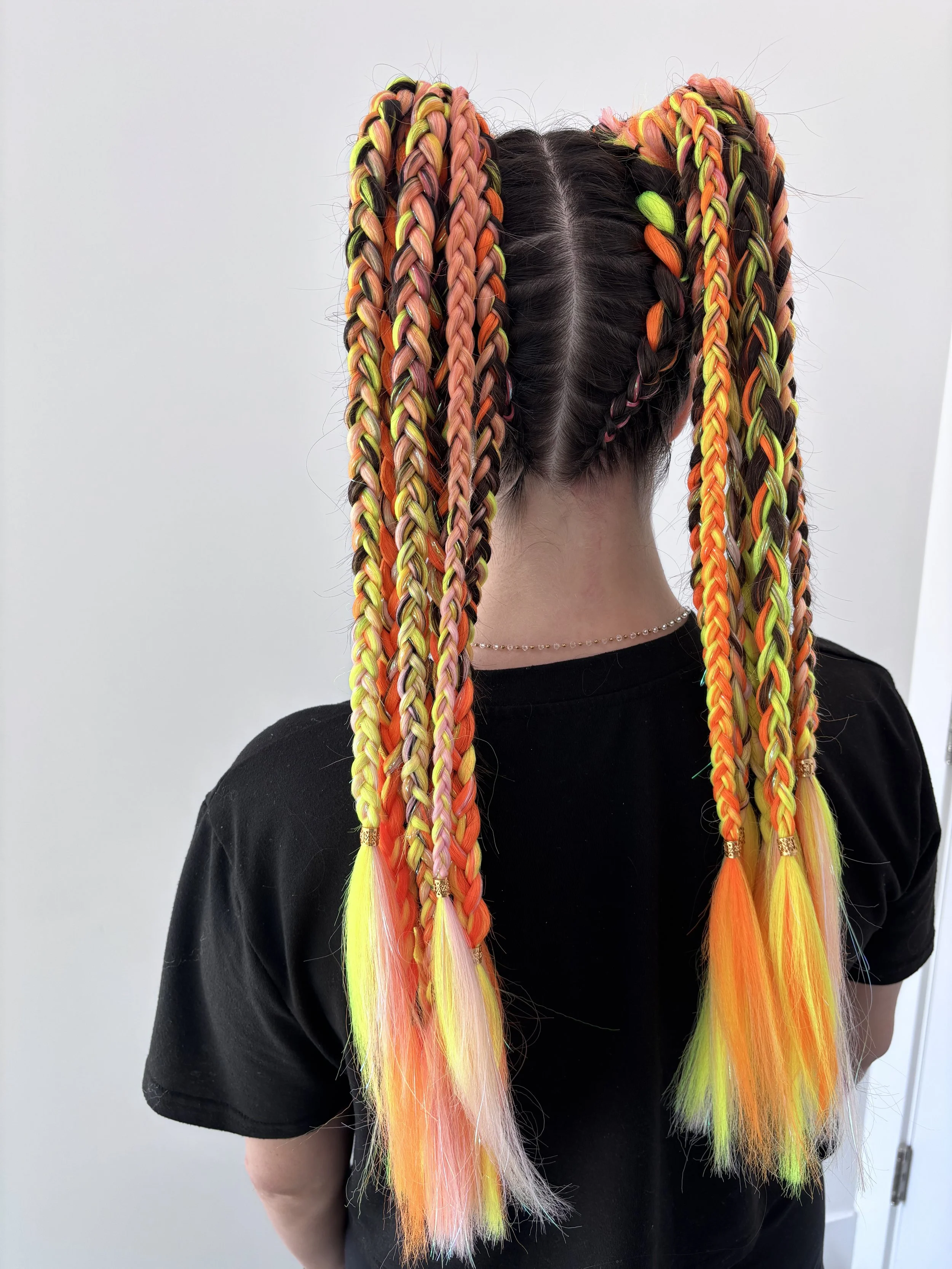 A person with multicolored box braids in shades of pink, orange, yellow, green, and black, styled into two high pigtails with some added colorful hair extensions.