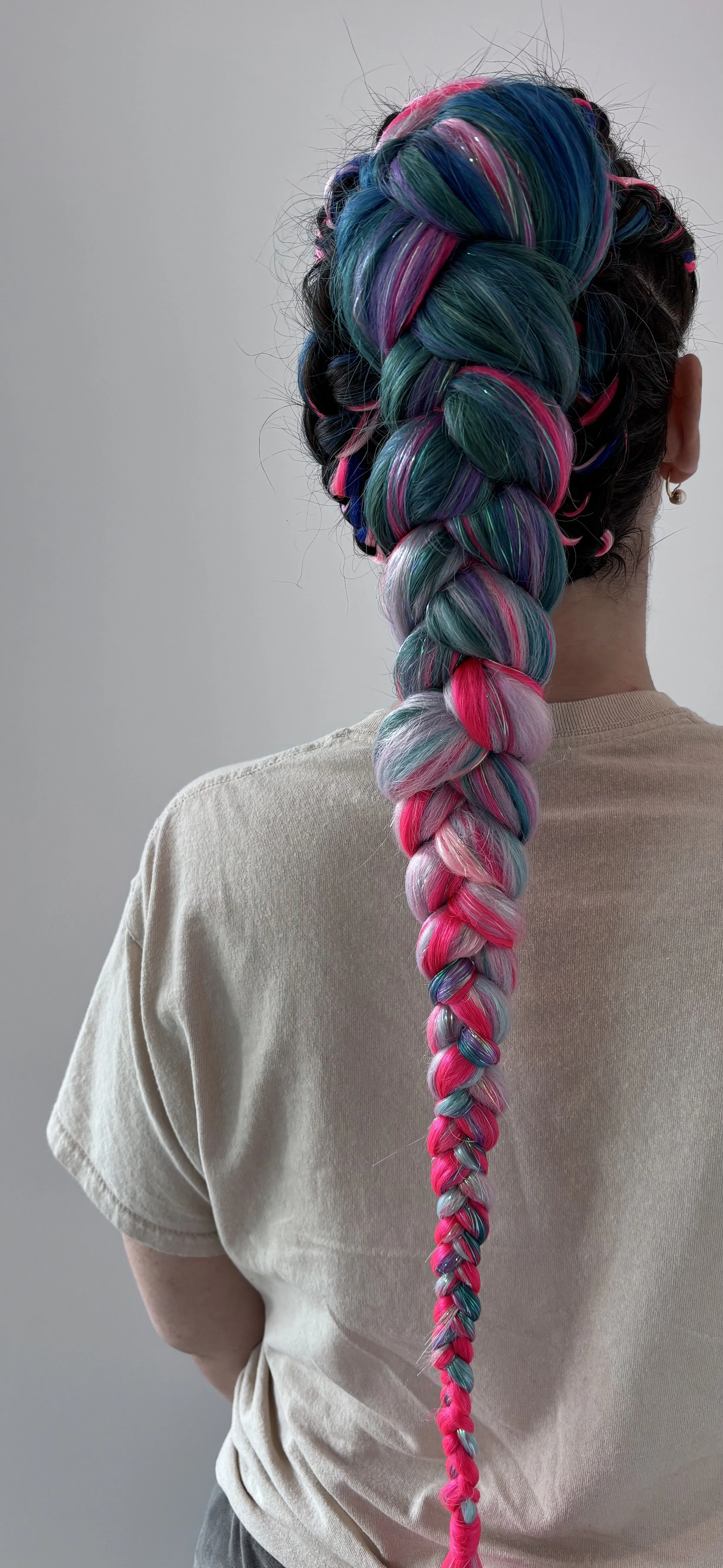 Person with long, colorful hair styled into a thick braid with shades of pink, blue, purple, green, and silver, standing against a plain light gray background.