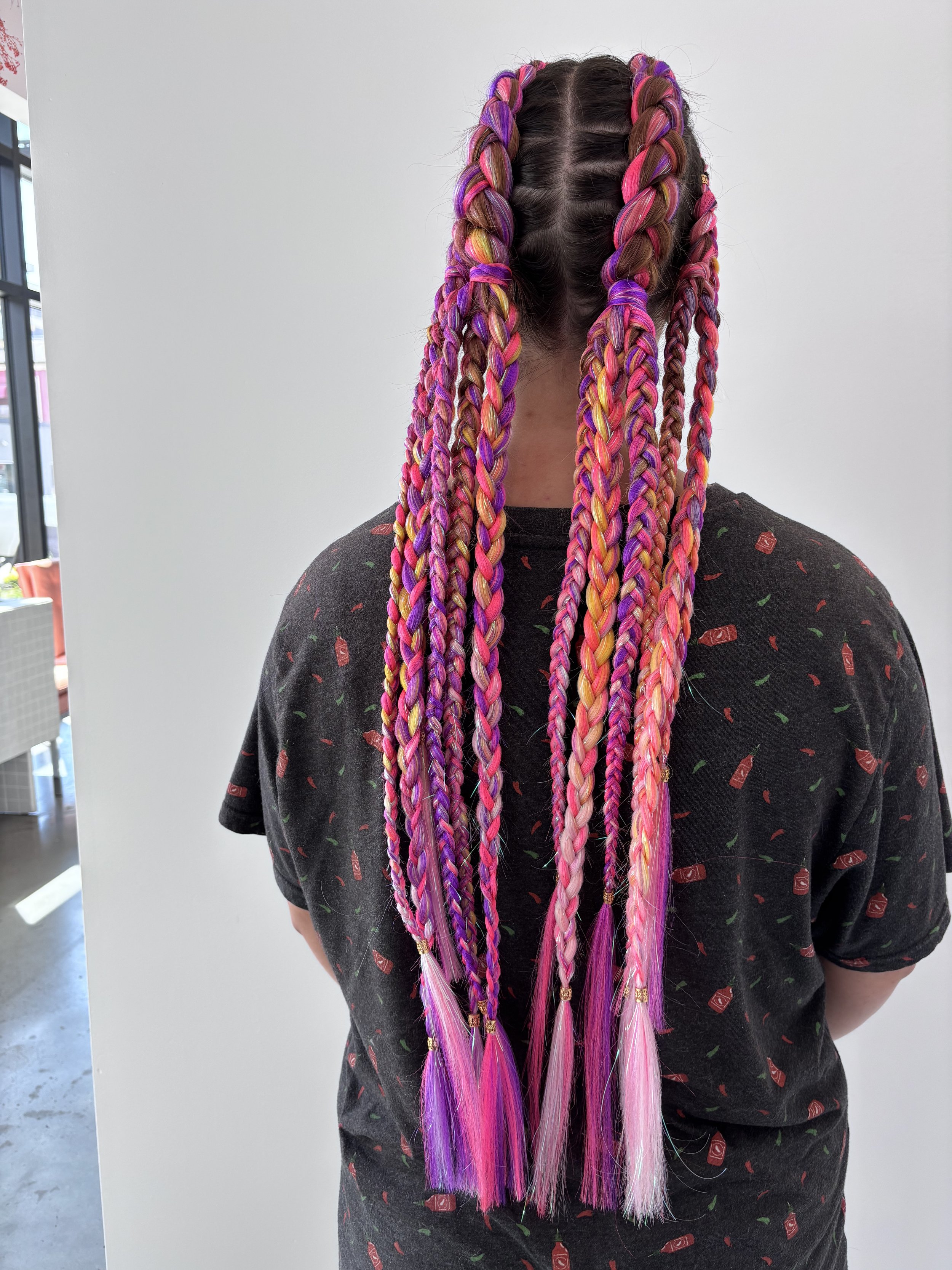 A person with long colorful braids in shades of purple, pink, yellow, and orange, standing in front of a plain wall, wearing a dark shirt with small red and green patterns.