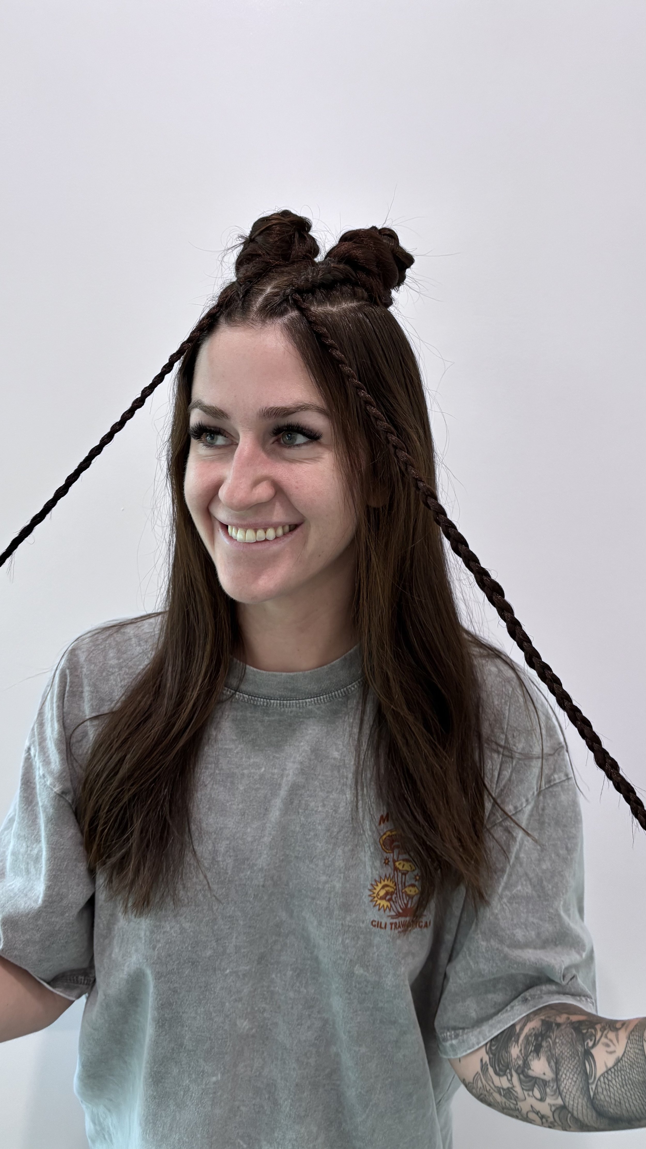 A young woman with long brown hair and tattoos on her right arm smiling while posing with her hair styled in two buns and braids against a plain white background.