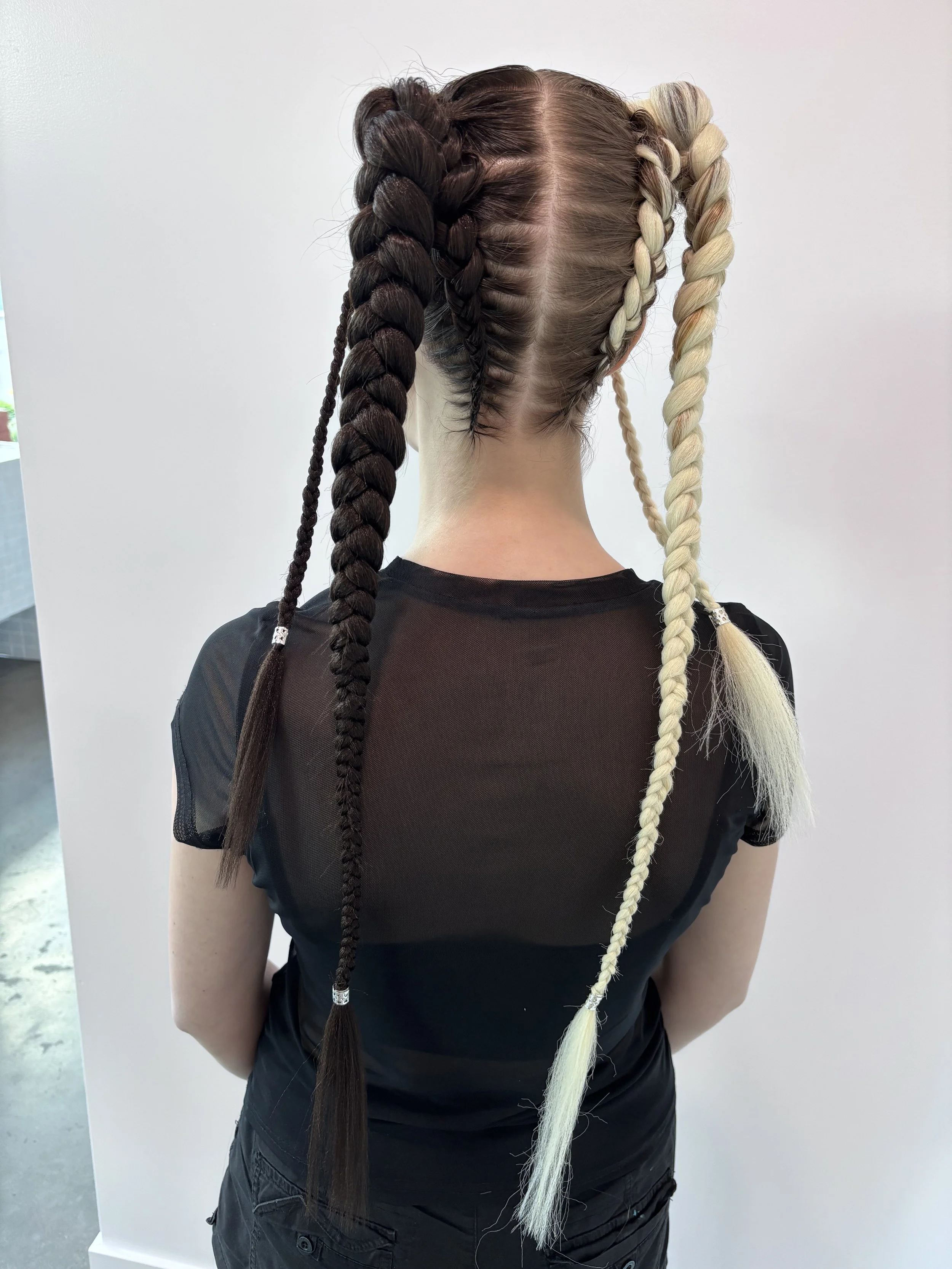 Back view of a woman with hair styled in four large braids, two dark brown and two blonde, decorated with small metallic beads at the ends.