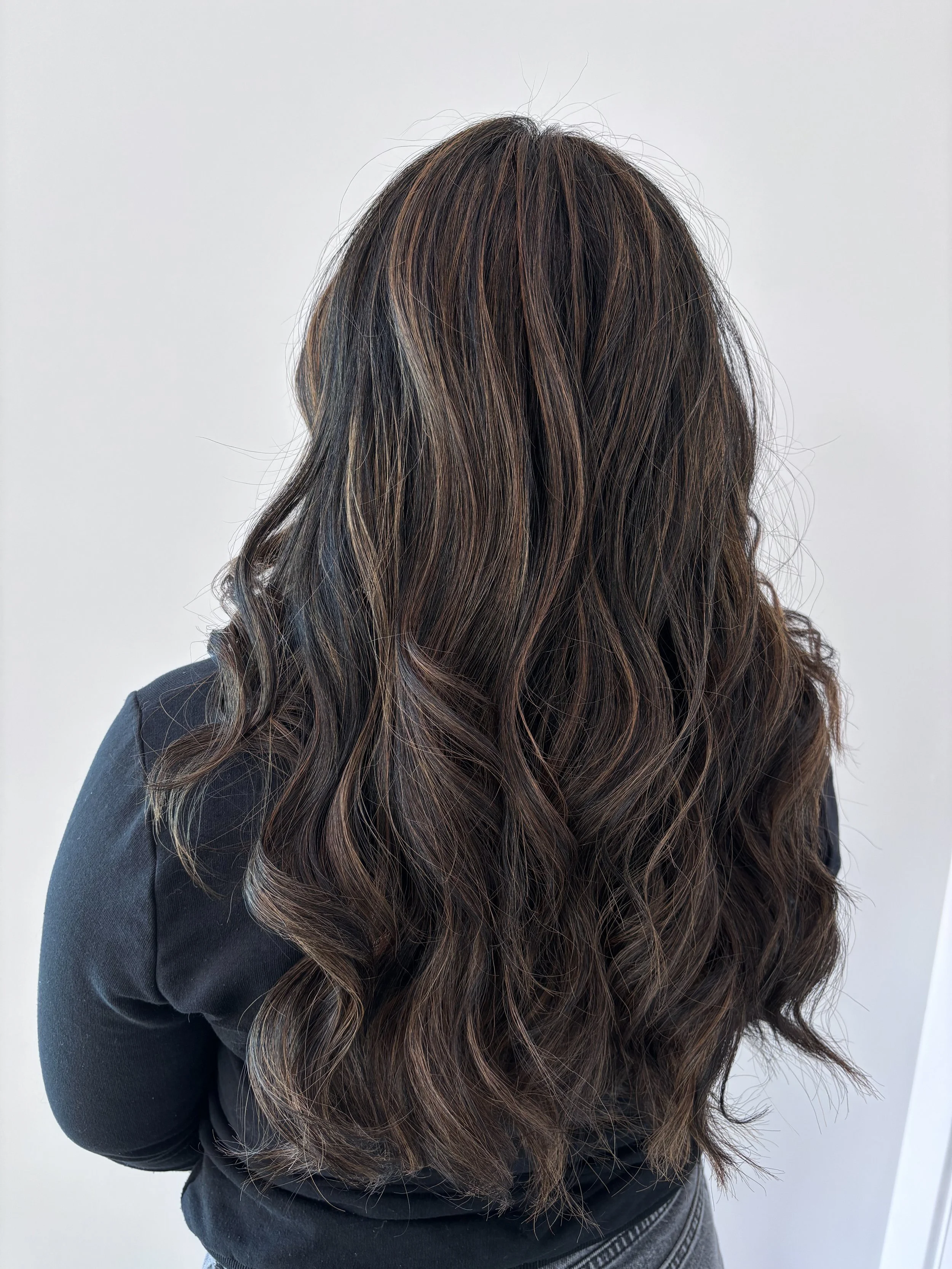 Back view of a woman with long, wavy brown hair with subtle highlights, dressed in a black top.