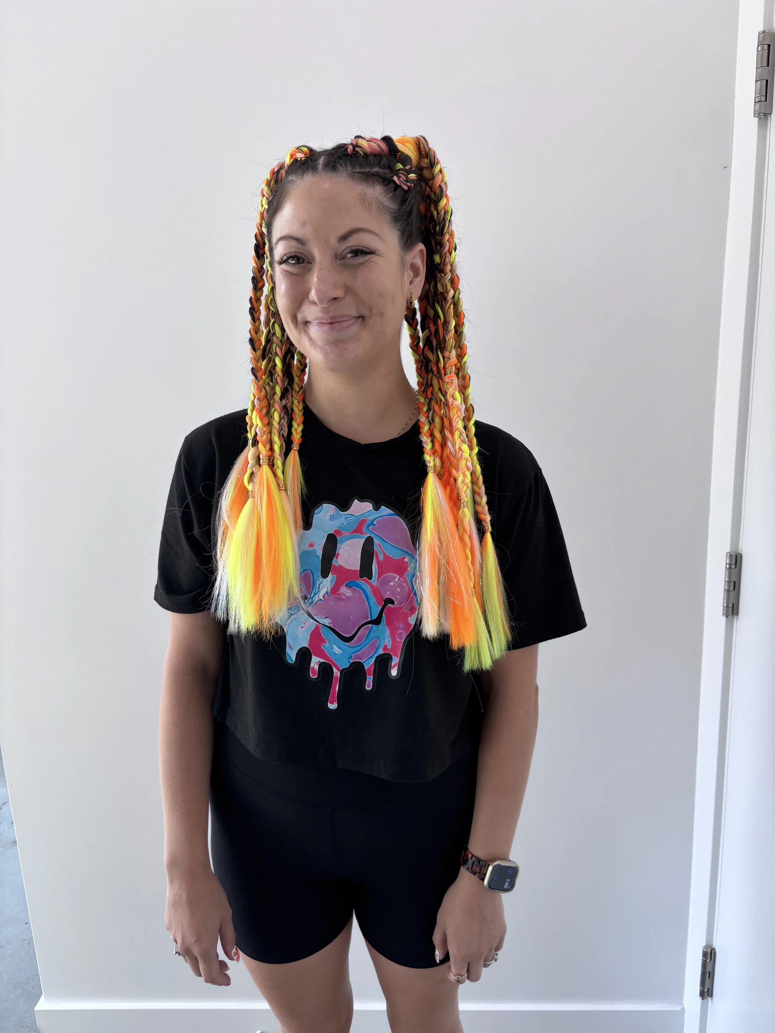 A woman with colorful braided hair and a big smile, wearing a black T-shirt with a colorful skull design, black shorts, a watch, and rings, standing in front of a plain white wall.