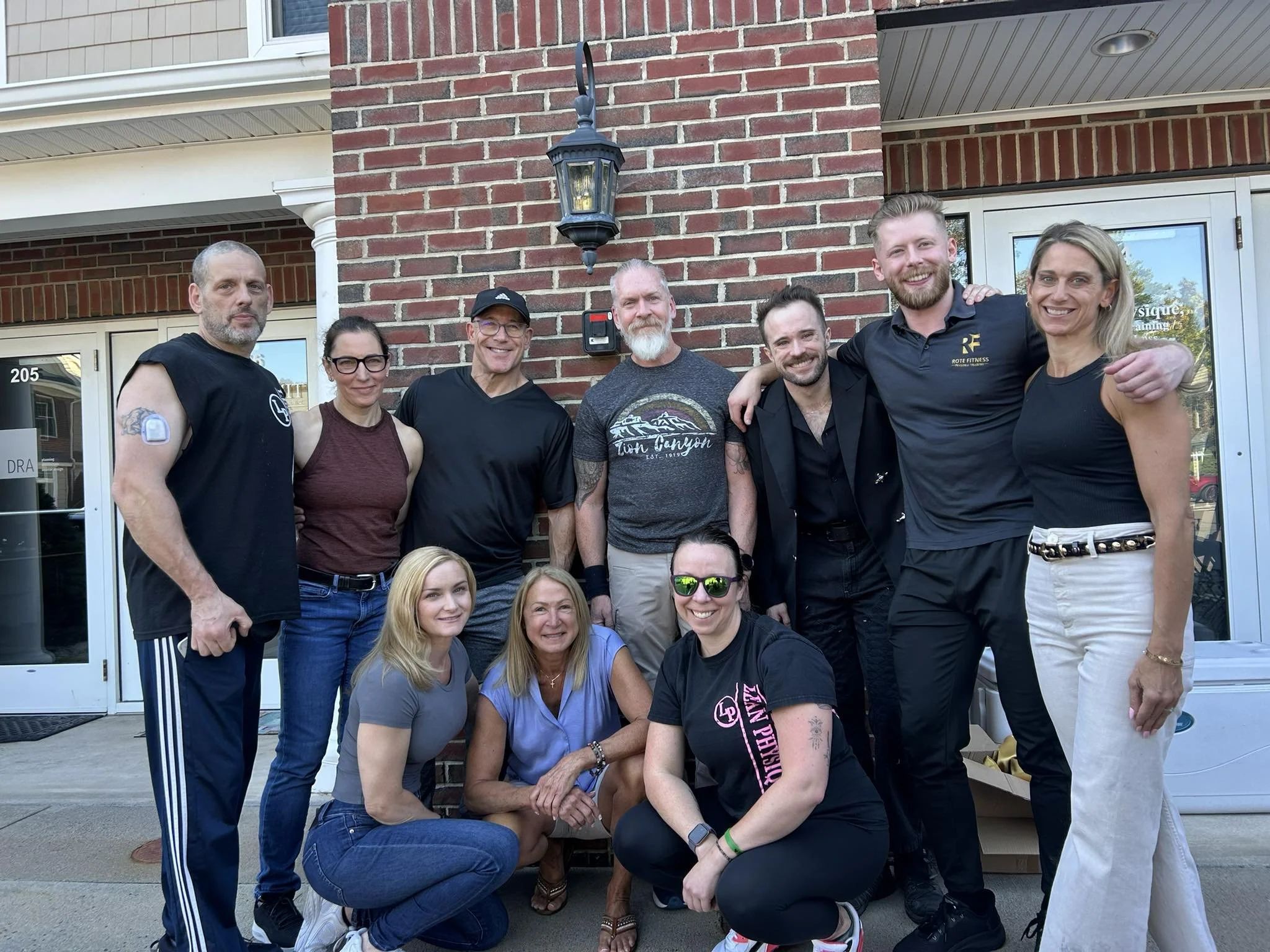 Group of ten people posing outside a building with a brick wall and glass door, some standing and some kneeling, all smiling.