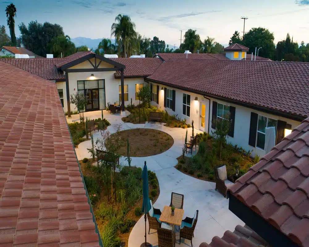 Nighttime view of a courtyard with benches, table with chairs, a closed umbrella, lush landscaping, and surrounding buildings with red-tiled roofs and exterior lighting.