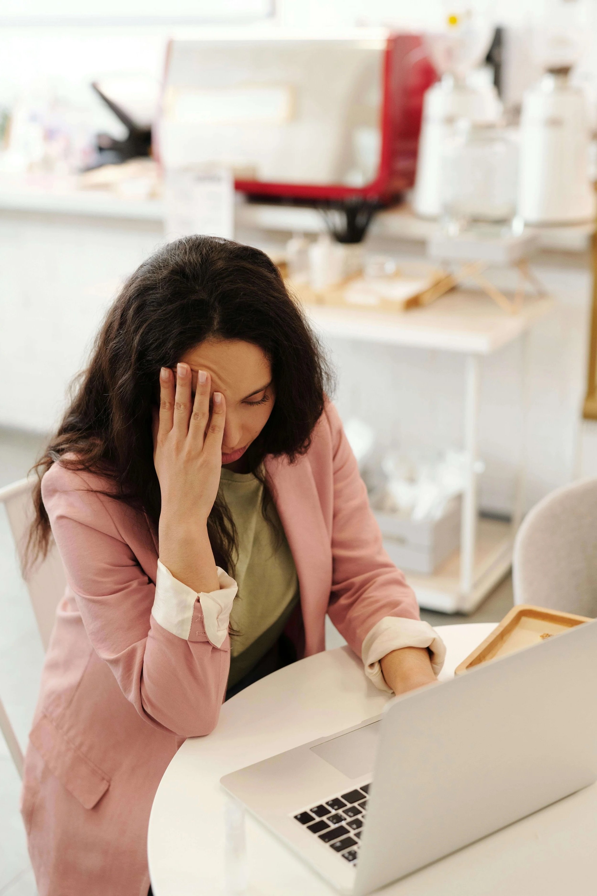 A woman in a pink jacket looking at a computer with her hand to her head, and a frustrated look on her face.