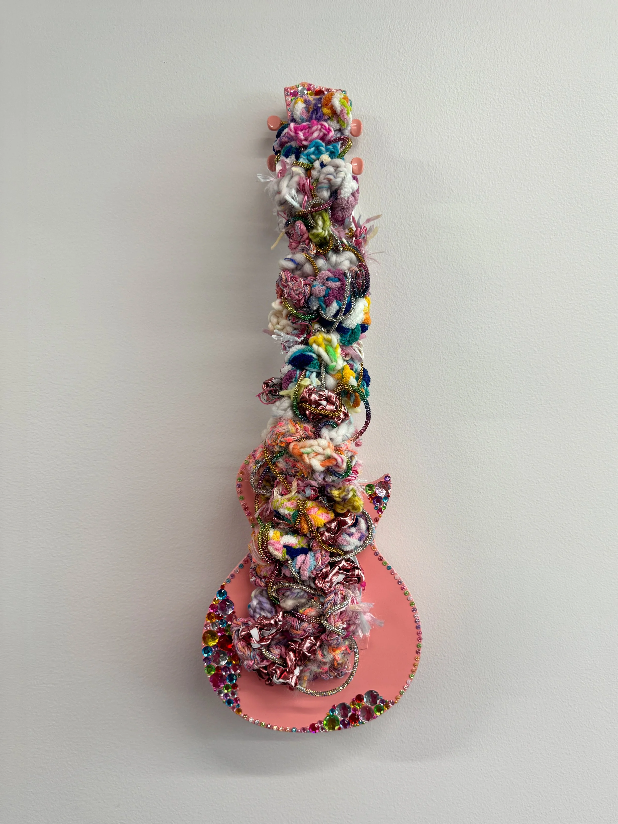 A fiber art piece comprised of interwoven fibers set within the strings of a ukulele, accented with applied rhinestones
