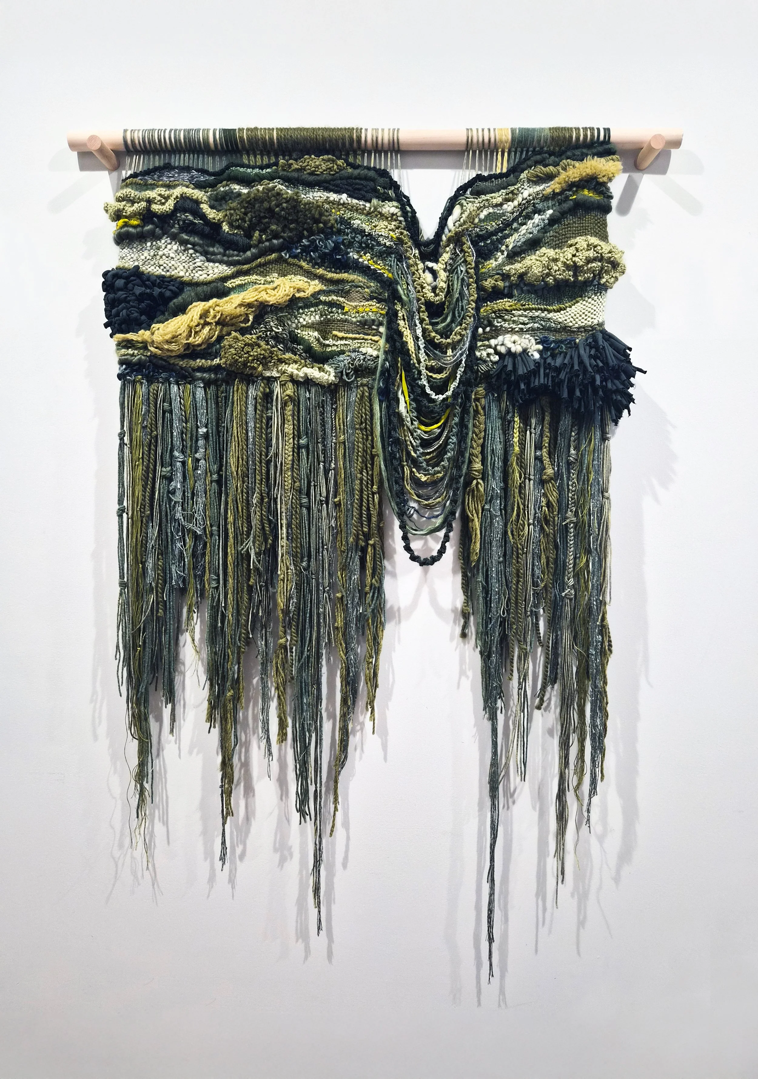 A fiber art piece comprised of interwoven fibers suspended from a wooden dowel