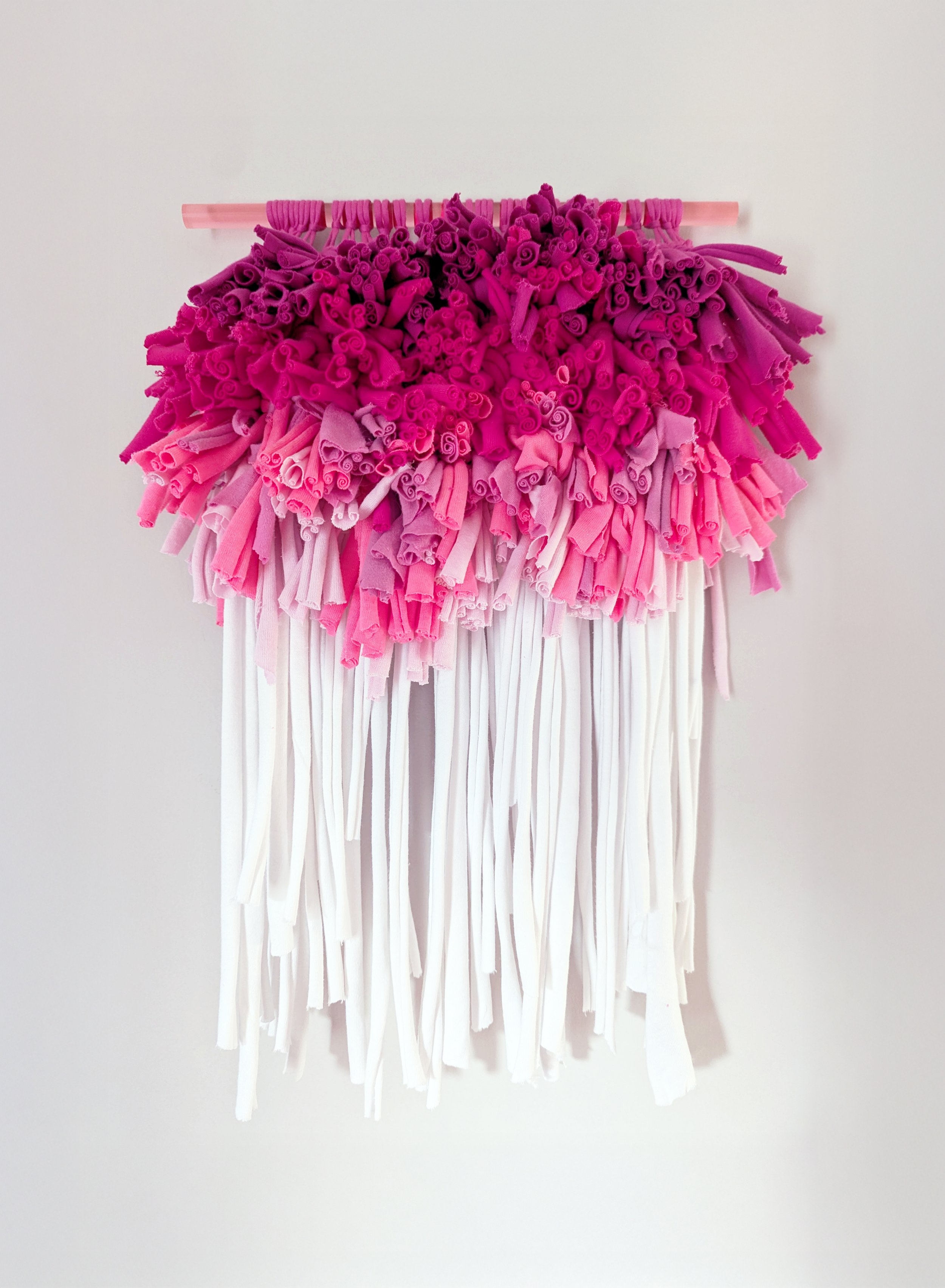 A fiber art piece comprised of interwoven T-shirts suspended from a translucent acrylic dowel
