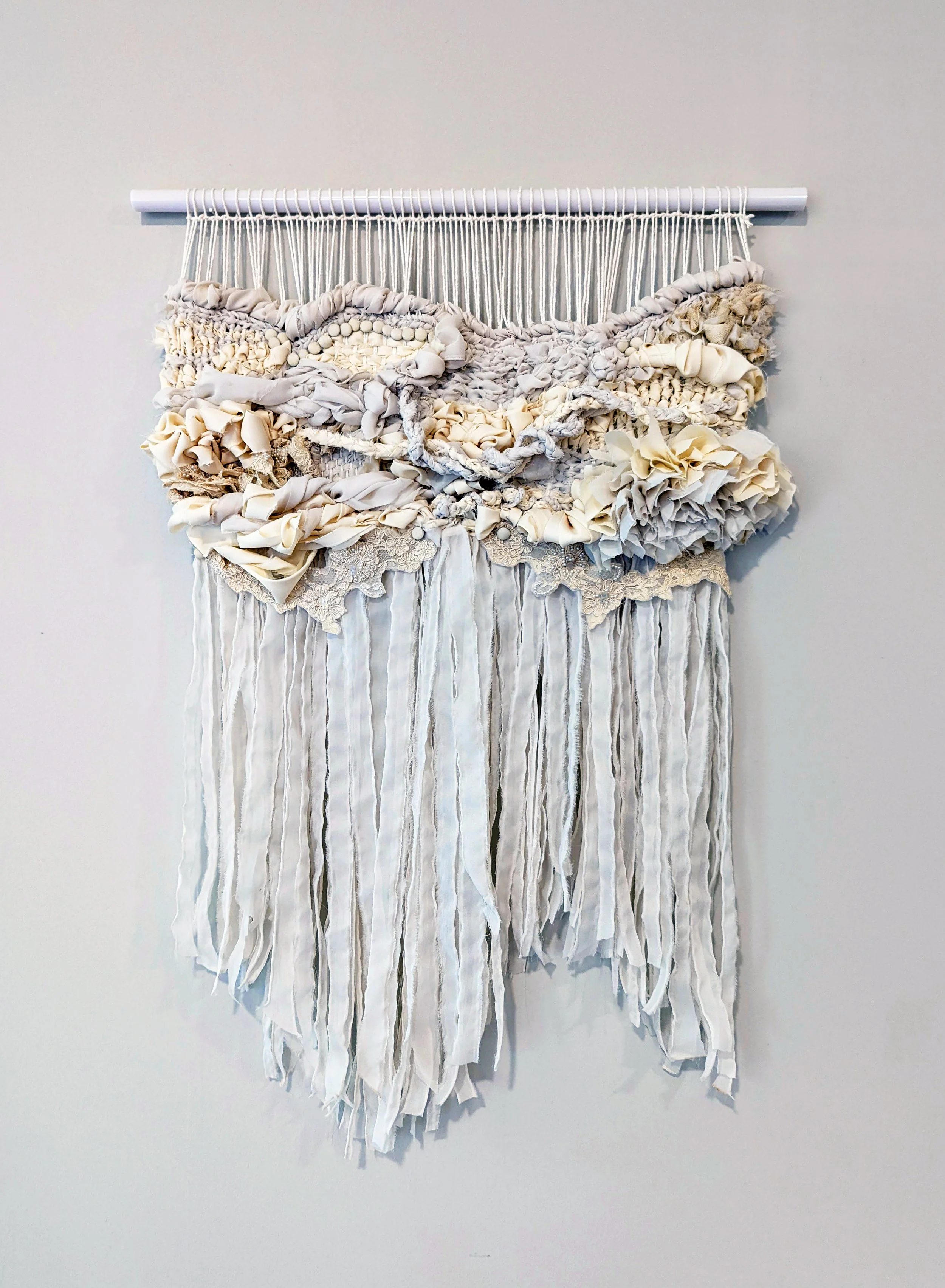 A fiber art piece comprised of interwoven reclaimed wedding dress remnants, lace, and buttons suspended from an acrylic dowel