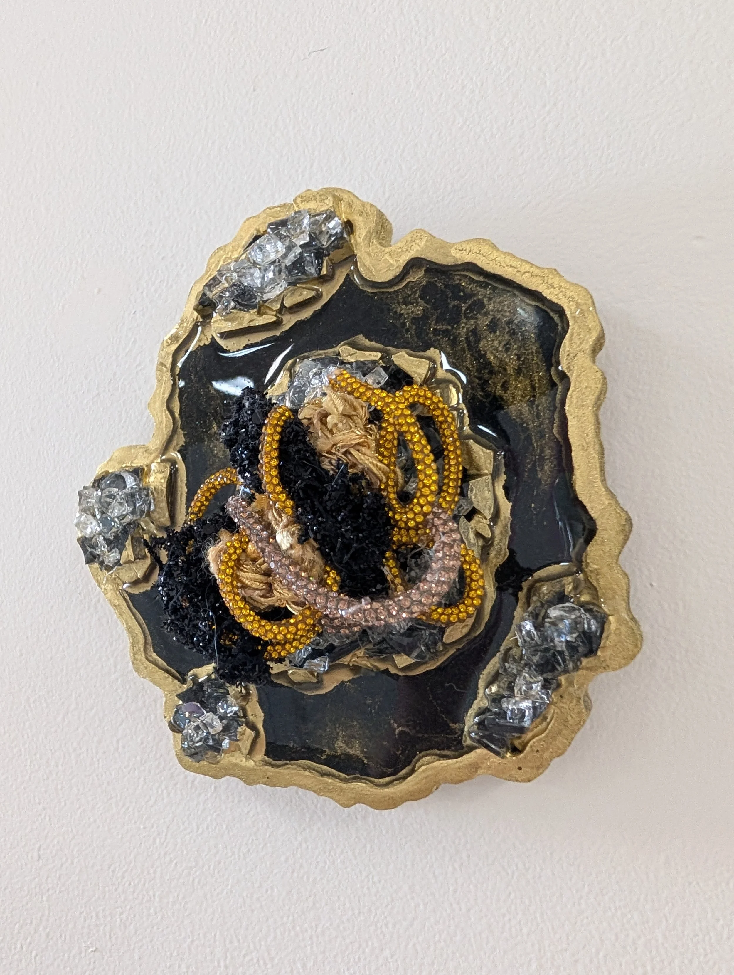 A fiber art piece comprised of interwoven fibers embedded in a resin geode with quartz, crushed glass, fire glass, mica powders, and glitter