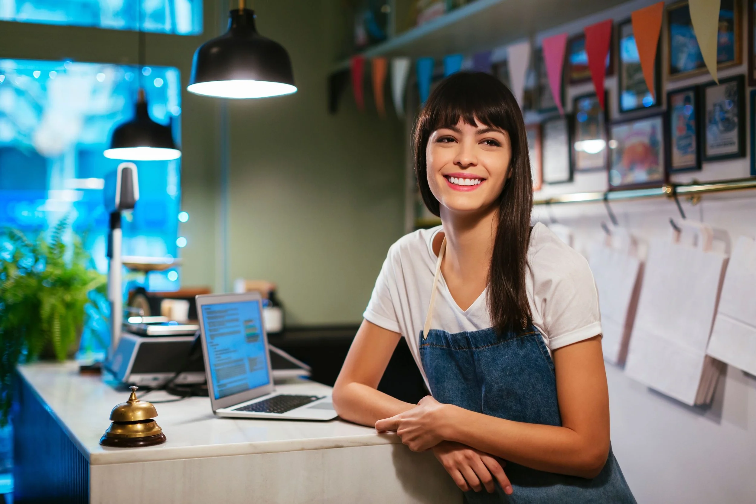 Smiling woman with dark hair, wearing a white t-shirt and denim apron, standing behind a reception counter in a cozy cafe or restaurant, with laptops and a small bell on the counter, colorful pennant banners hanging above and framed pictures on the wall in the background.