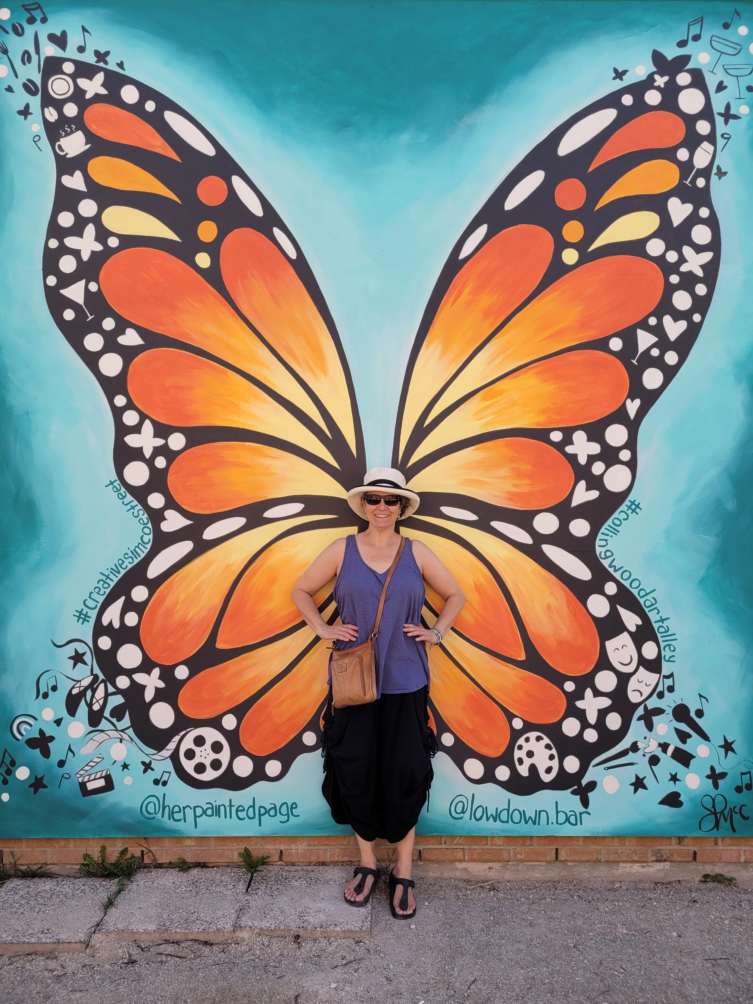 Dee Hutton, artist and maker, standing with hands on hips in front of large bright orange monarch butterfly wings in Collingwood, ON