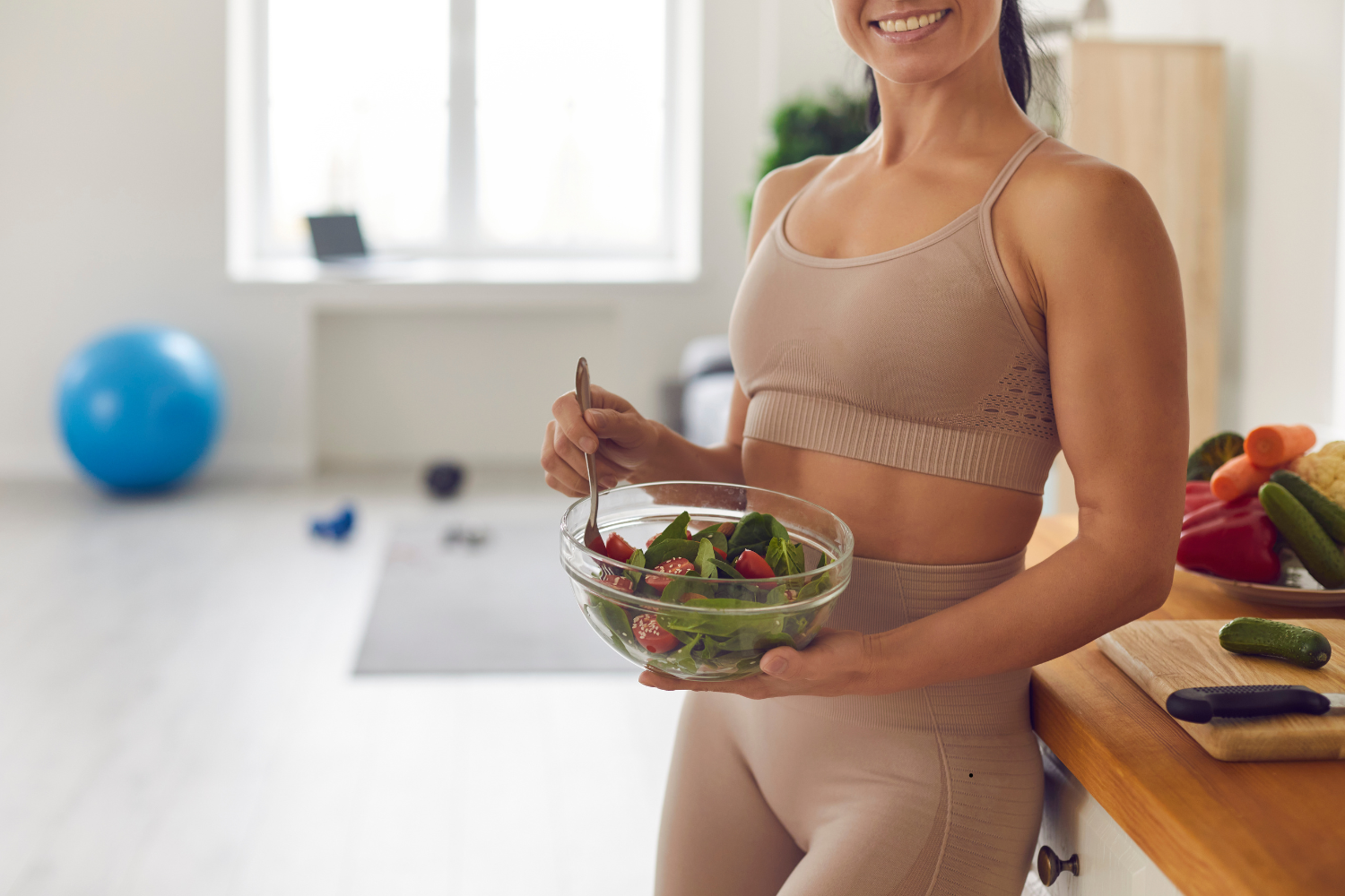Image is of a healthy woman in work out gear surrounded by work out equipment and vegetables and eating a salad