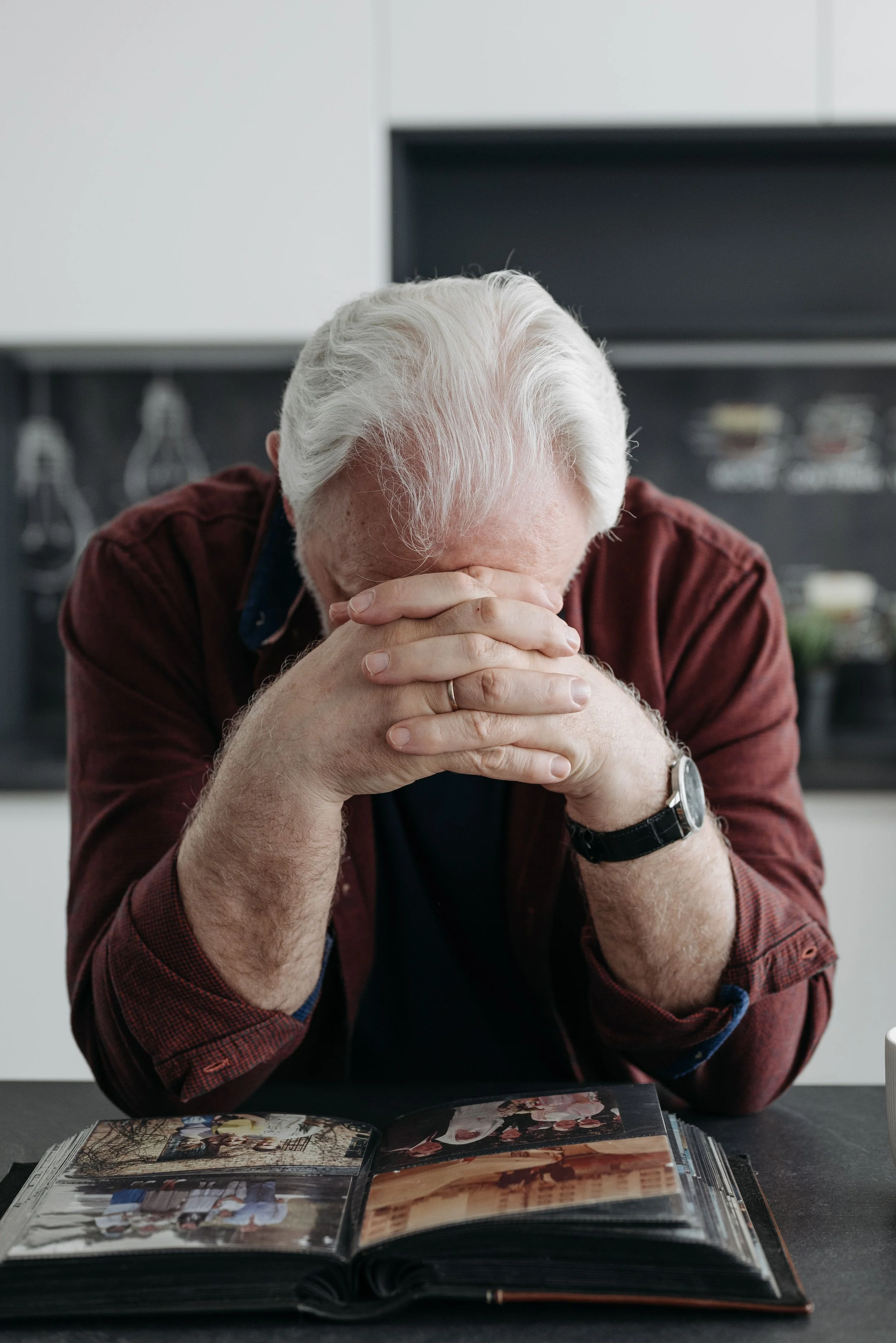 An older man with white hair sitting with his head lowered over an open photo album. Copy
