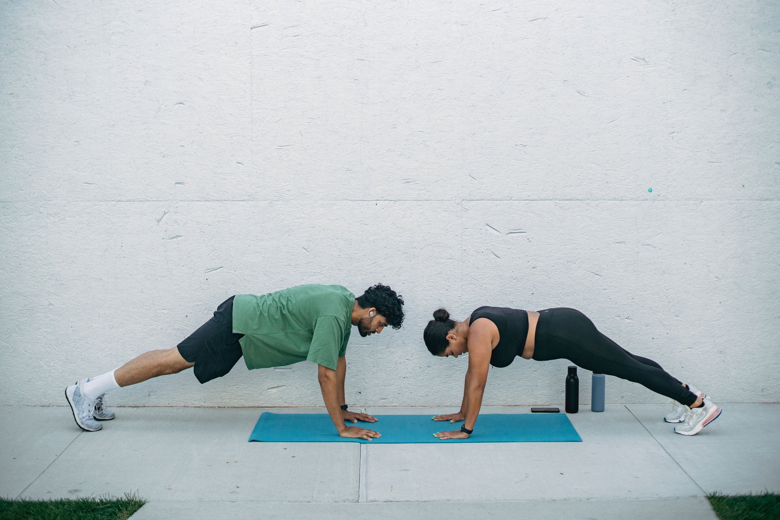 Two adults exercising on yoga mats in a plank position outdoors, facing each other. They are dressed in athletic clothing, with water bottles and a phone nearby against a light concrete wall.