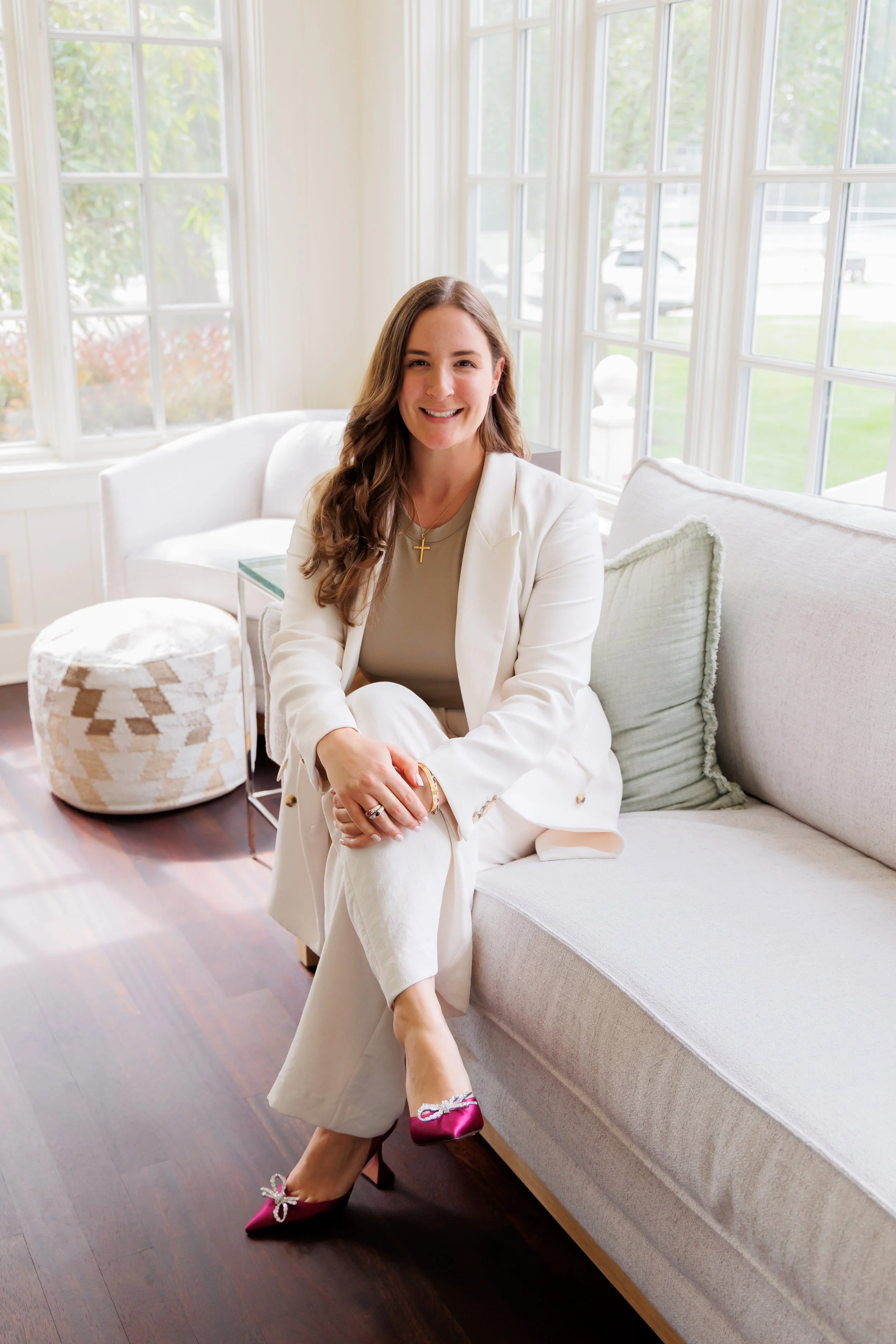 A woman sitting on a light-colored sofa in a bright room with large windows. She is wearing a white suit and magenta high heels. Her hands are clasped over her knee, and she is smiling at the camera.