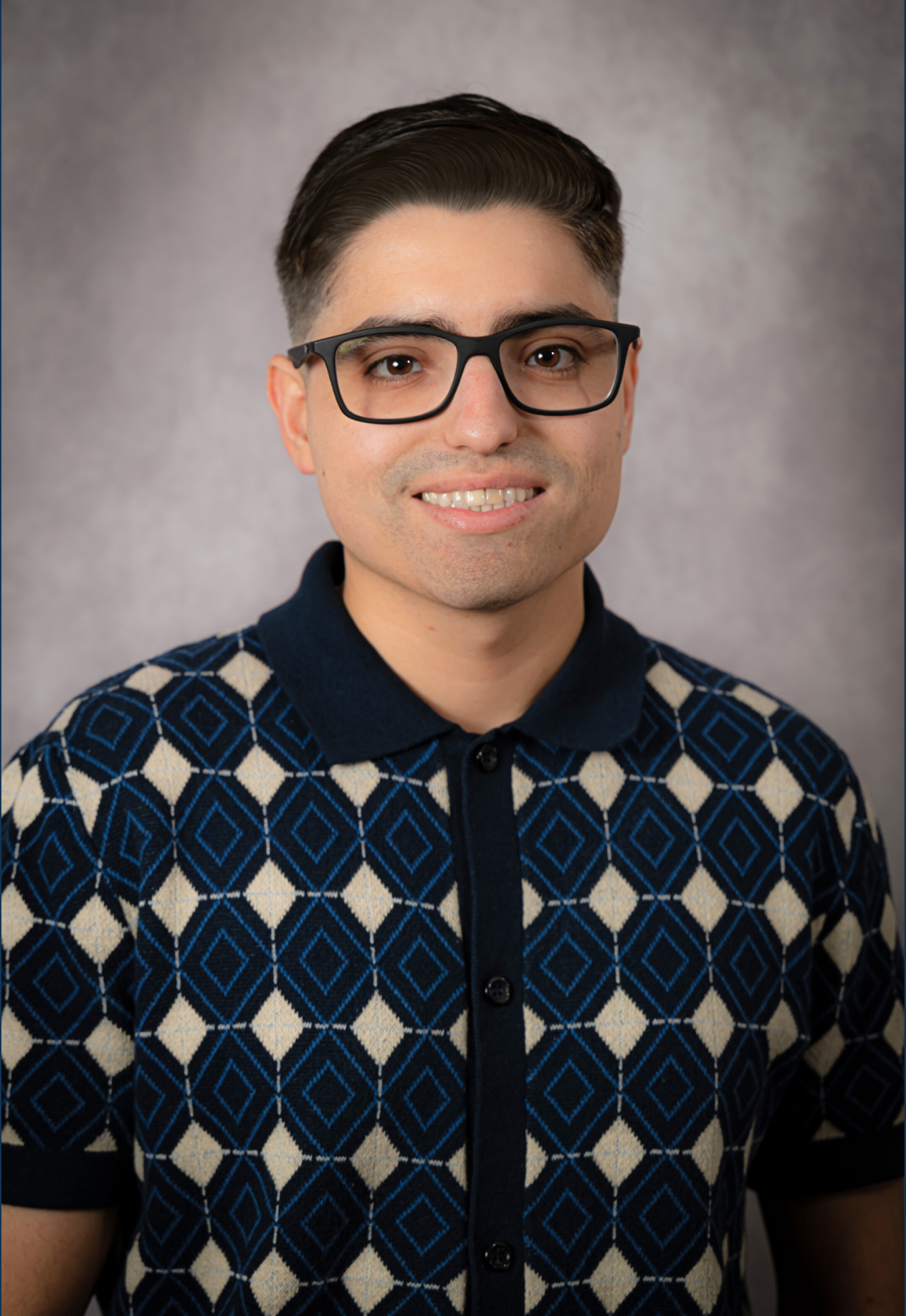 A young man with dark hair and glasses wearing a patterned blue and beige polo shirt, smiling at the camera against a gray background.