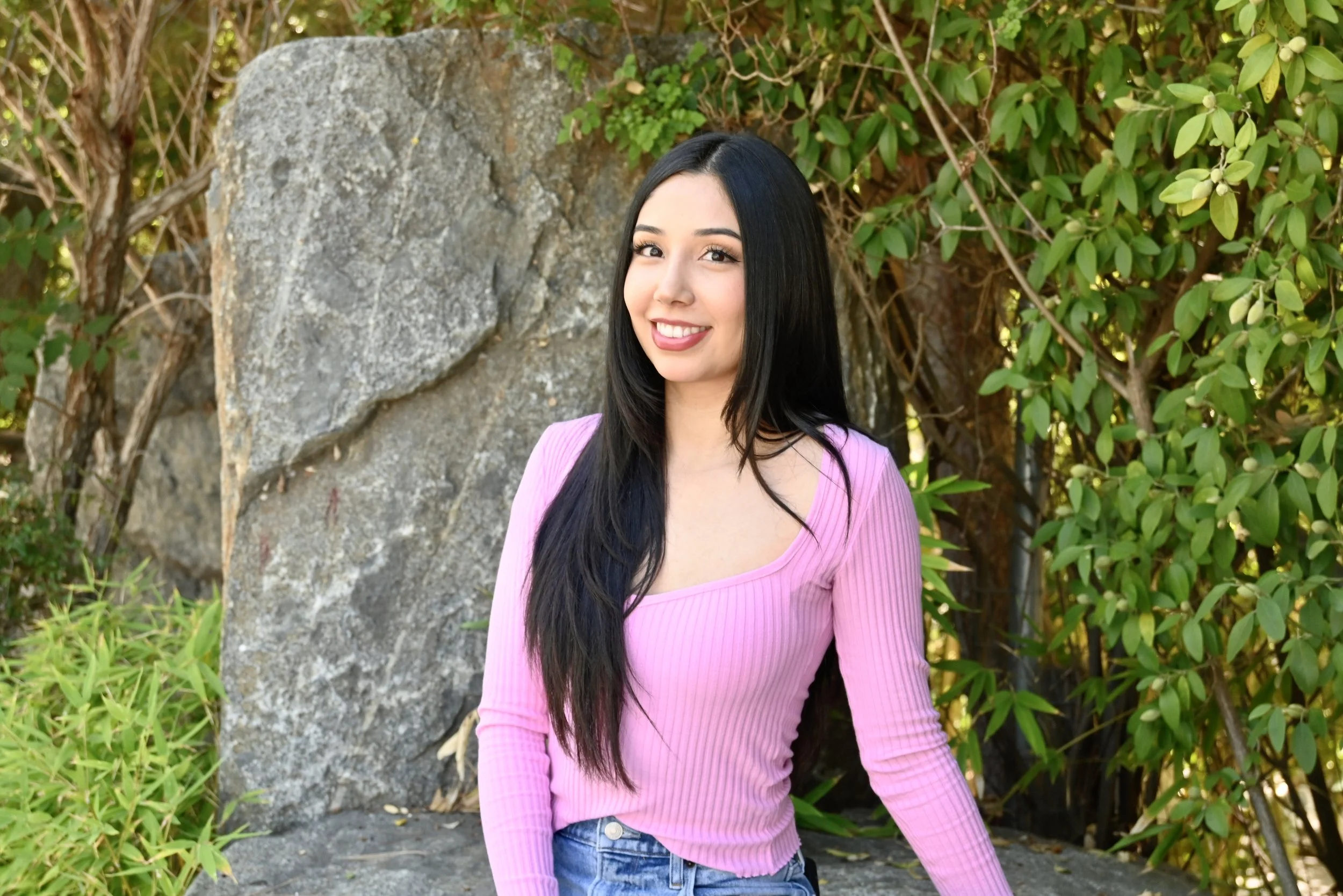 A young woman with long black hair, wearing a pink long-sleeve top and blue jeans, standing outdoors in front of a large rock and green foliage, smiling at the camera.