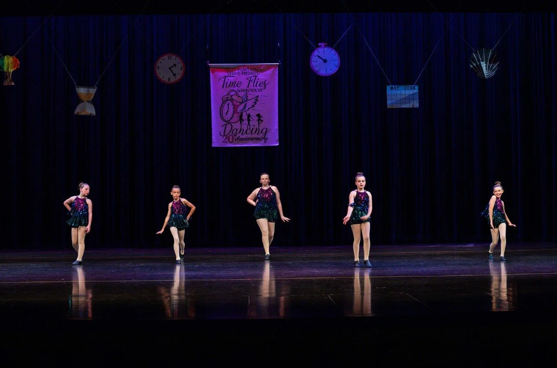 Five young dancers in sparkly purple and green costumes performing a jazz routine on stage at Crazy Moves Dance Studio’s 20th anniversary recital under a “Time Flies” banner..jpg