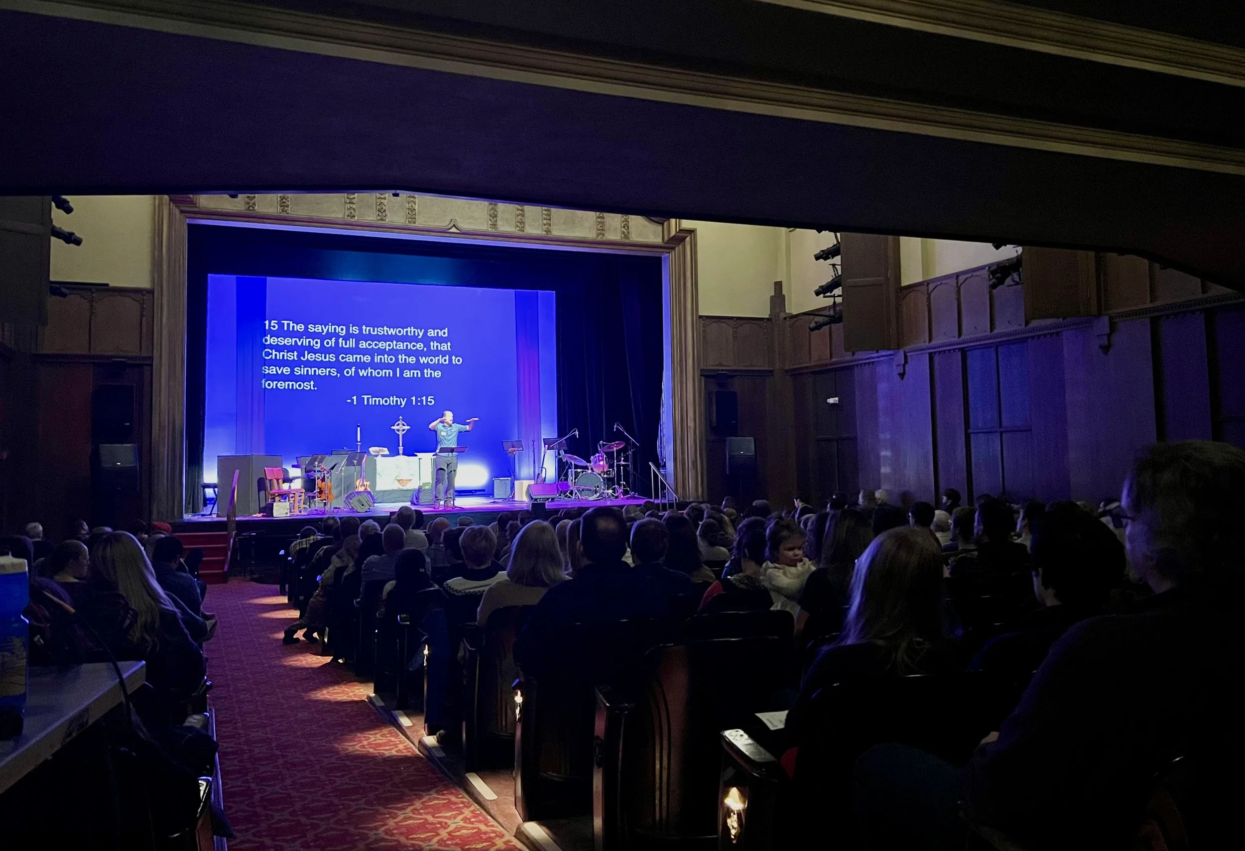 An audience watches a church service proceeding on stage in a theatre. Verses are projected on to the wall behind the pastor.