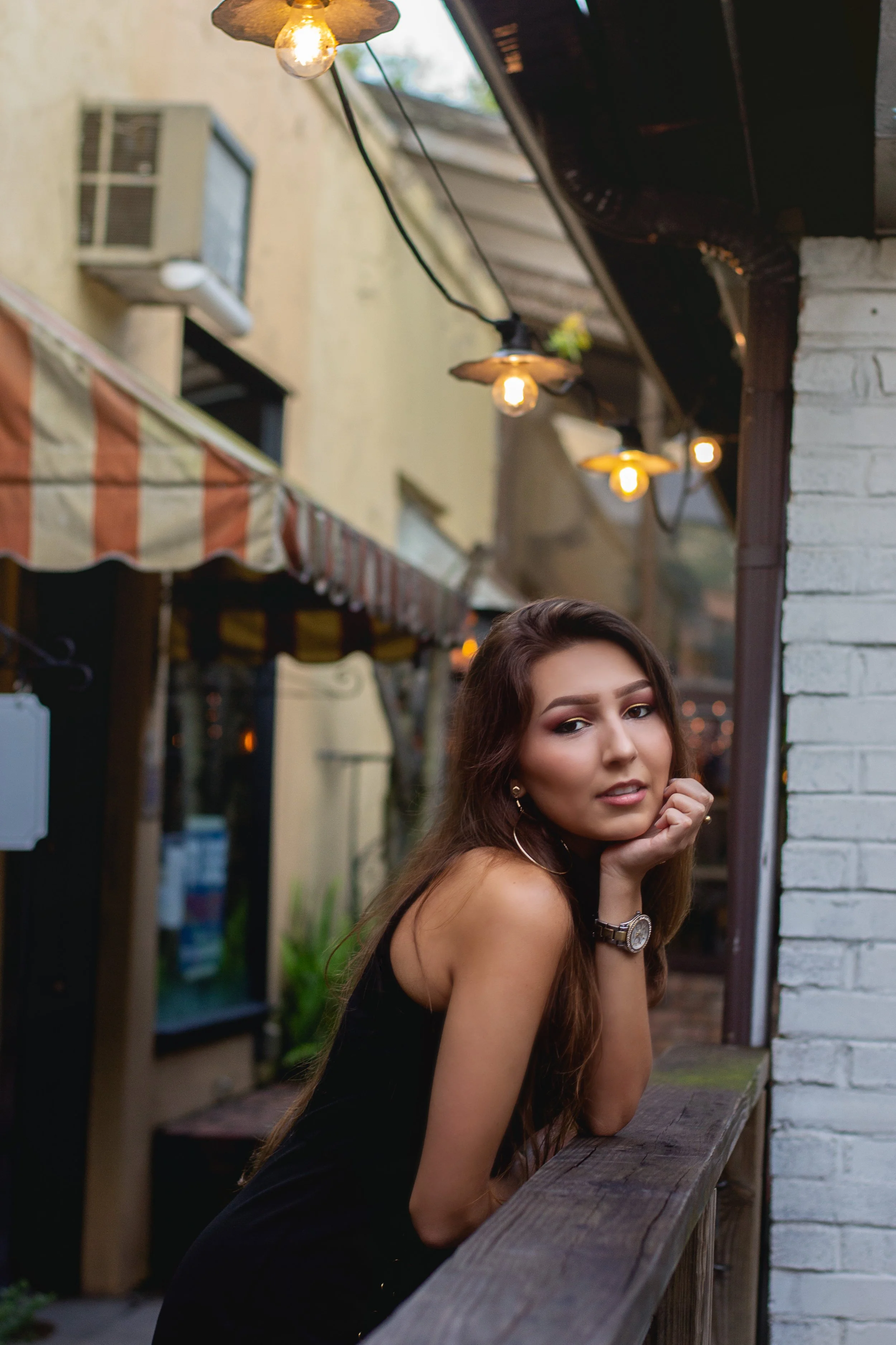 A young woman with long brown hair in a black top leans on a wooden railing outdoors at dusk, with string lights hanging overhead and a building with an awning and an air conditioning unit in the background.