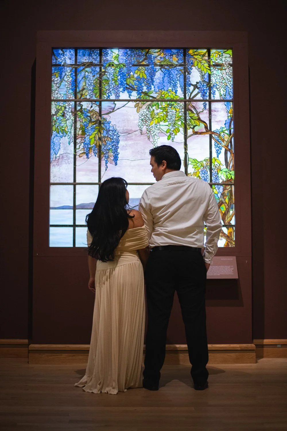 A couple stands close together in front of a large stained glass window depicting blue wisteria flowers and green leaves, with a scenic landscape in the background.