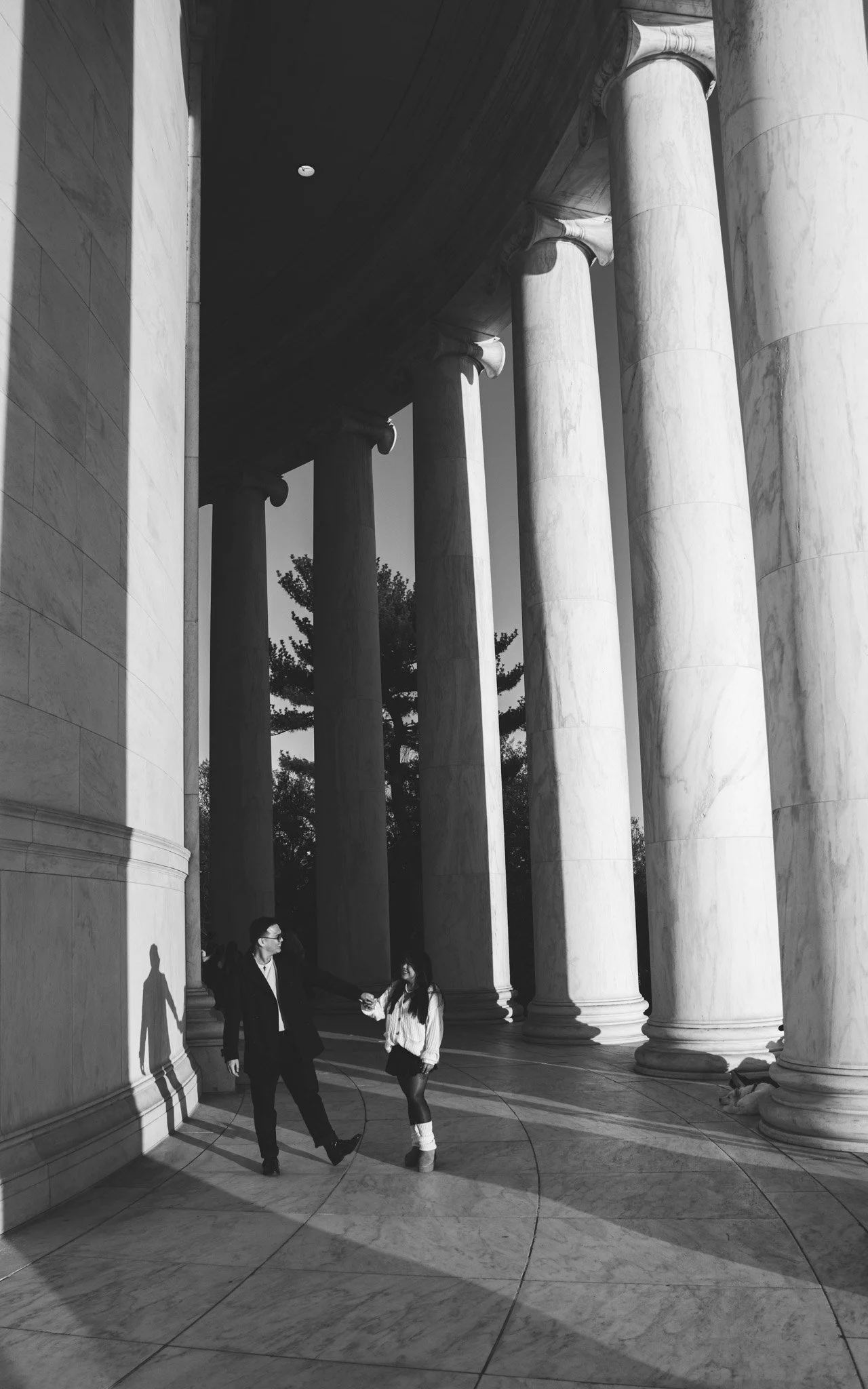 A black and white photo of two people holding hands and walking near large marble columns of a classical building, with their shadows cast on the floor.
