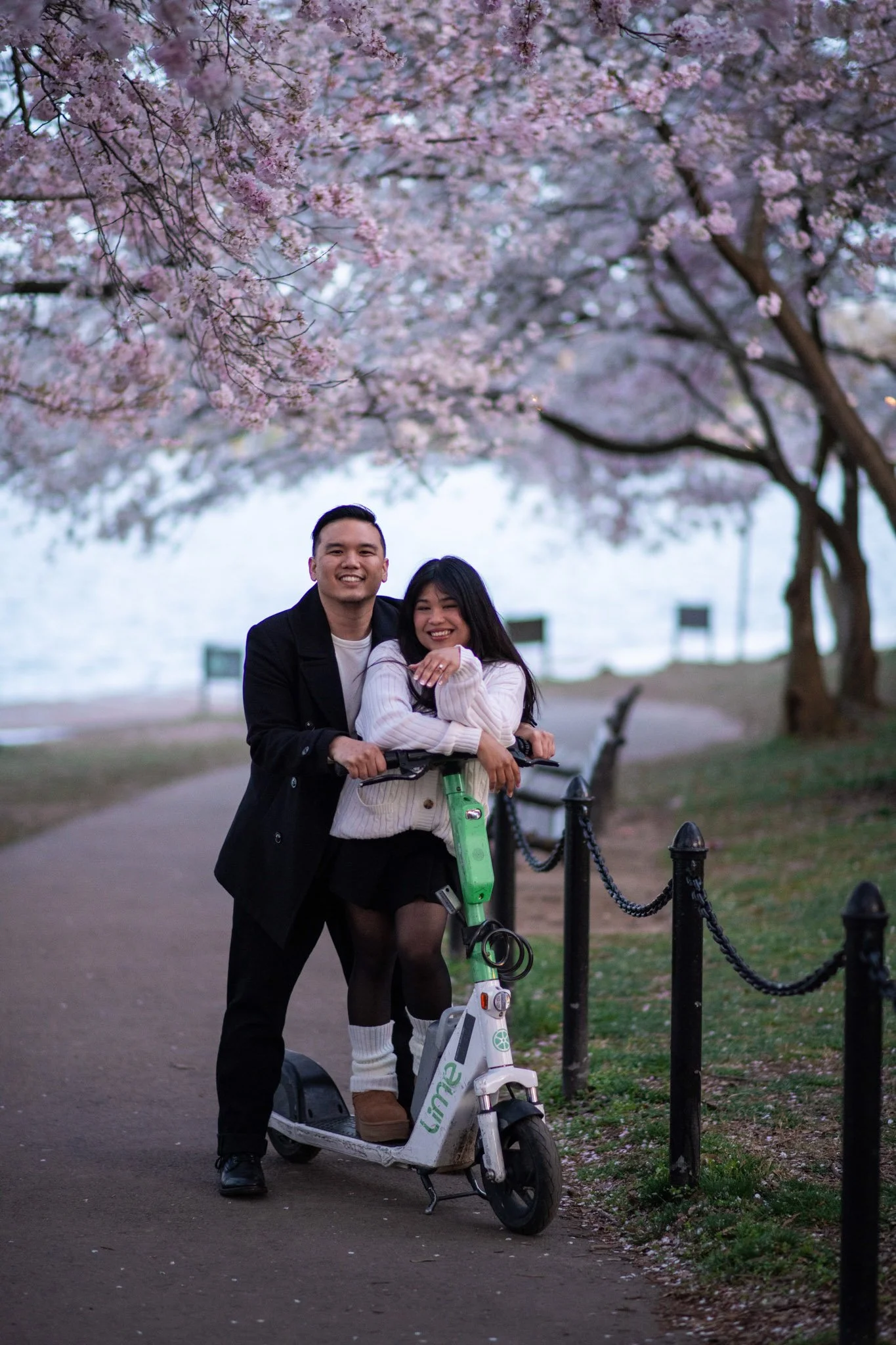 A couple riding a green electric scooter together under blooming cherry blossom trees in a park.