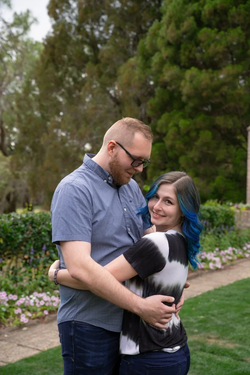 A couple hugging outdoors in a garden with trees and flowers in the background.