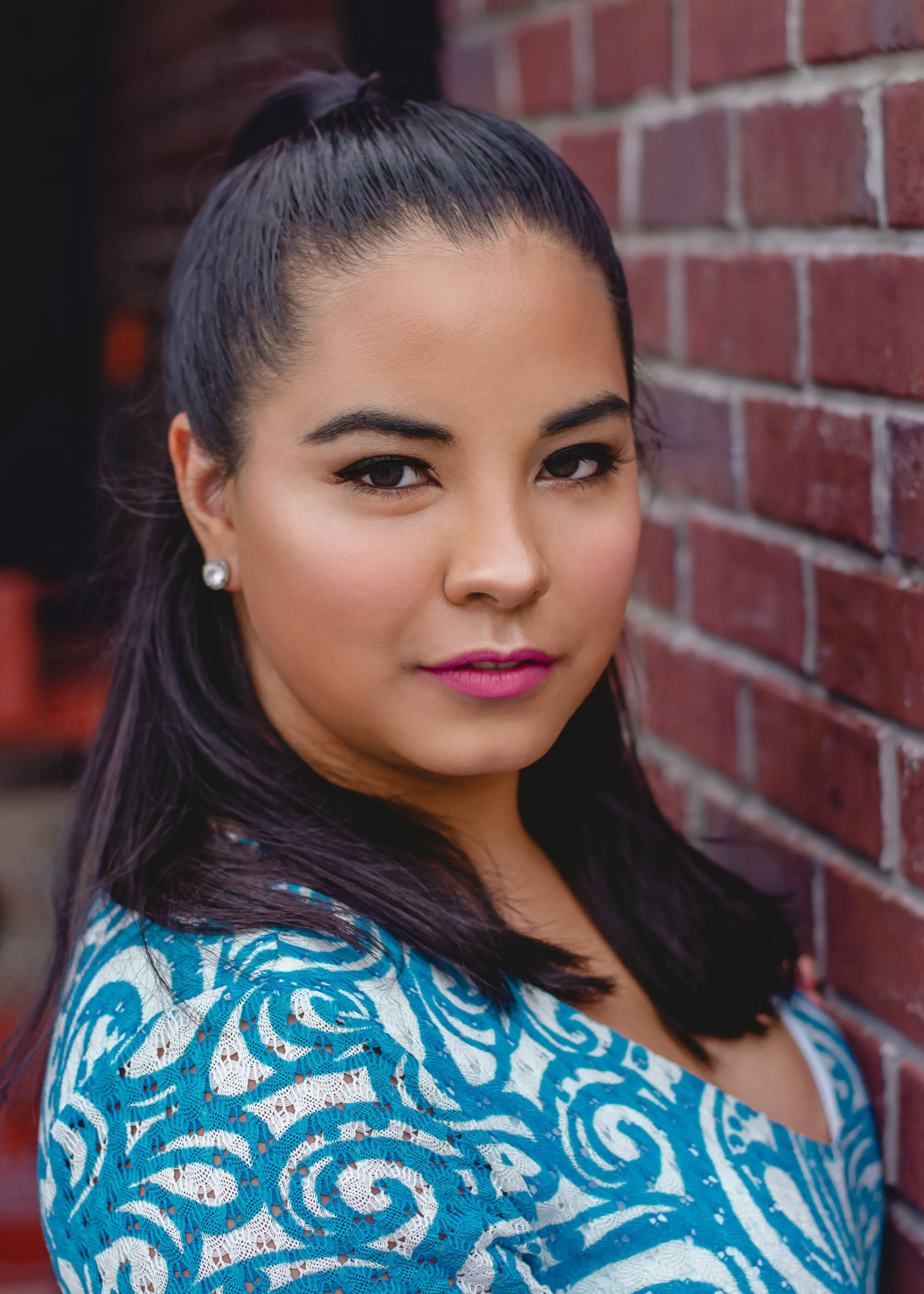 A woman with dark hair tied in a ponytail, wearing a blue and white patterned lace top, standing against a red brick wall.