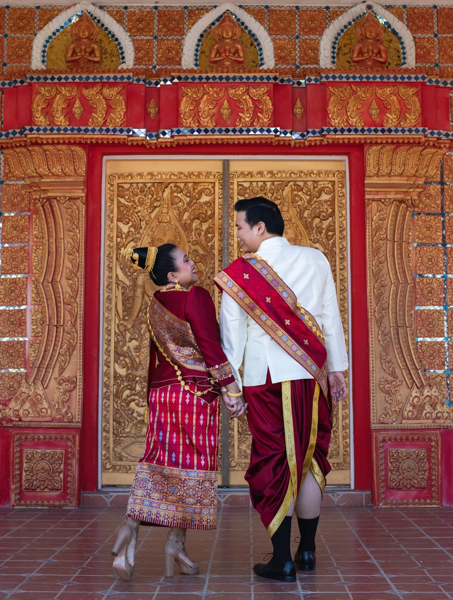A couple in traditional Thai wedding attire holding hands, facing each other in front of intricately carved wooden doors with gold and red accents, surrounded by ornate decorations and statues.