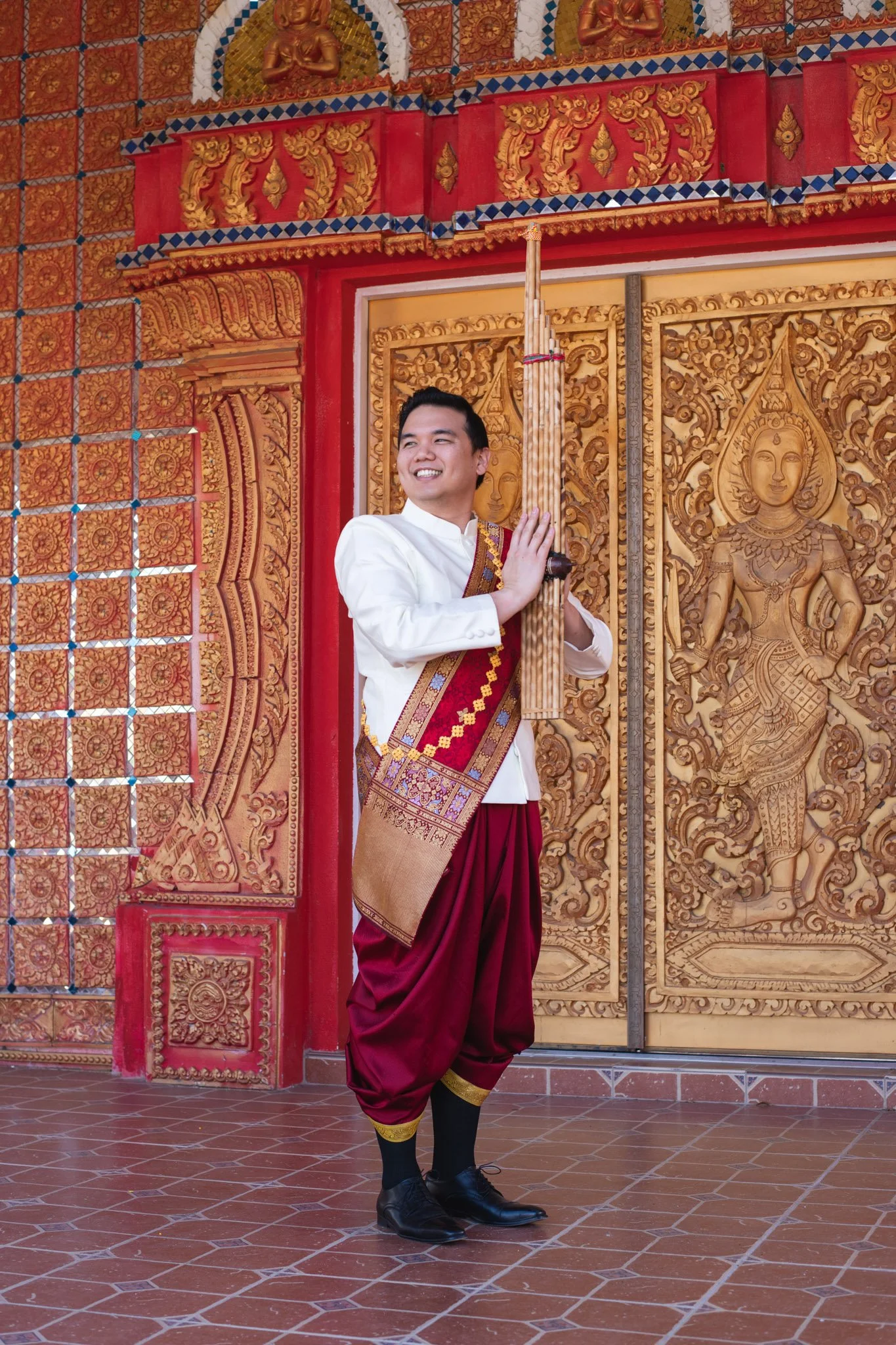 A man dressed in traditional Thai attire holding a musical instrument standing in front of intricately carved wooden doors at a Thai temple.
