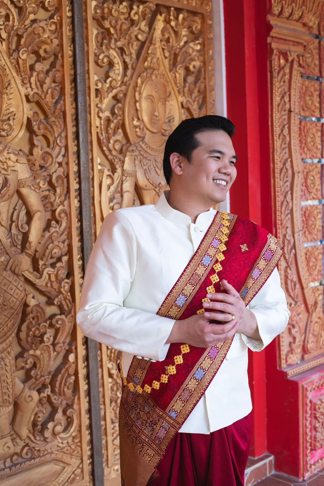 A young man dressed in traditional Thai attire stands in front of intricately carved wooden doors with Buddhist and mythological figures, smiling and holding his hands in a traditional greeting.