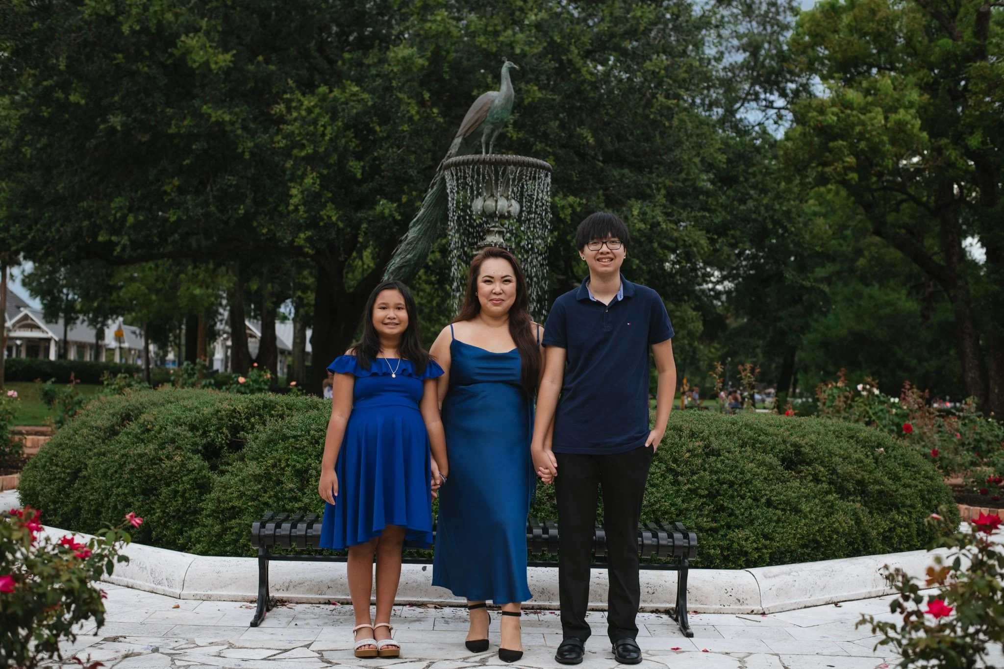 Three people standing in front of a fountain with a peacock statue, surrounded by greenery and bushes, during daytime.